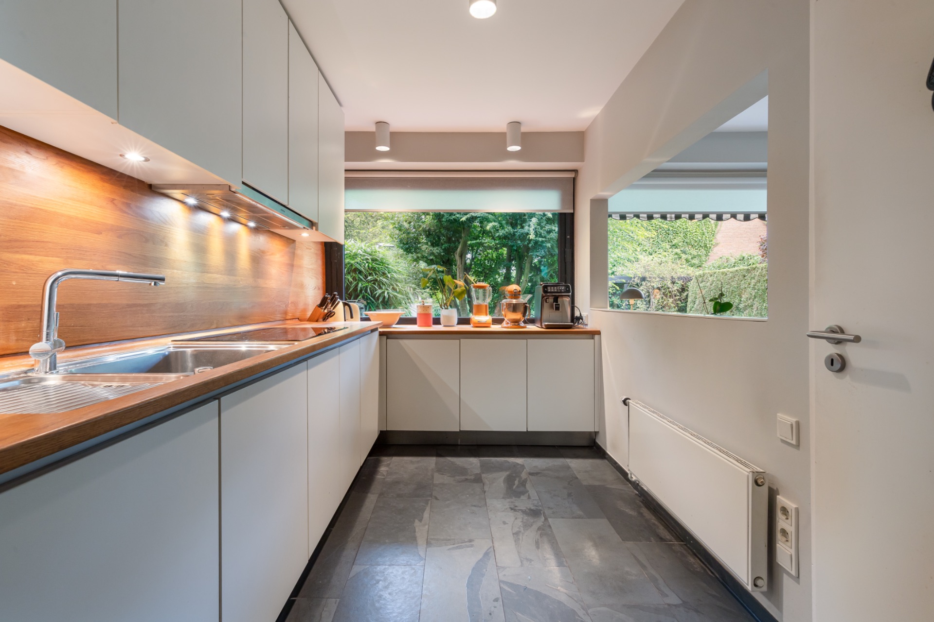 Moderne Küche mit weißen Schränken, Holzarbeitsplatte und großem Fenster zum Garten.