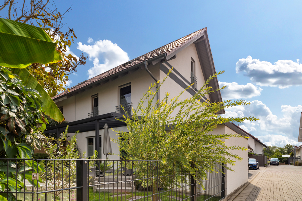 Modernes, weißes Wohnhaus mit Garten und Umzäunung an einer ruhigen Straße bei Sonnenschein.