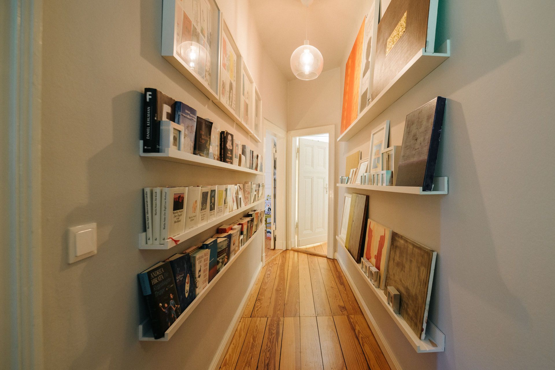 Schmaler Flur mit Holzboden, Bücherregalen und Bilderrahmen an den Wänden.