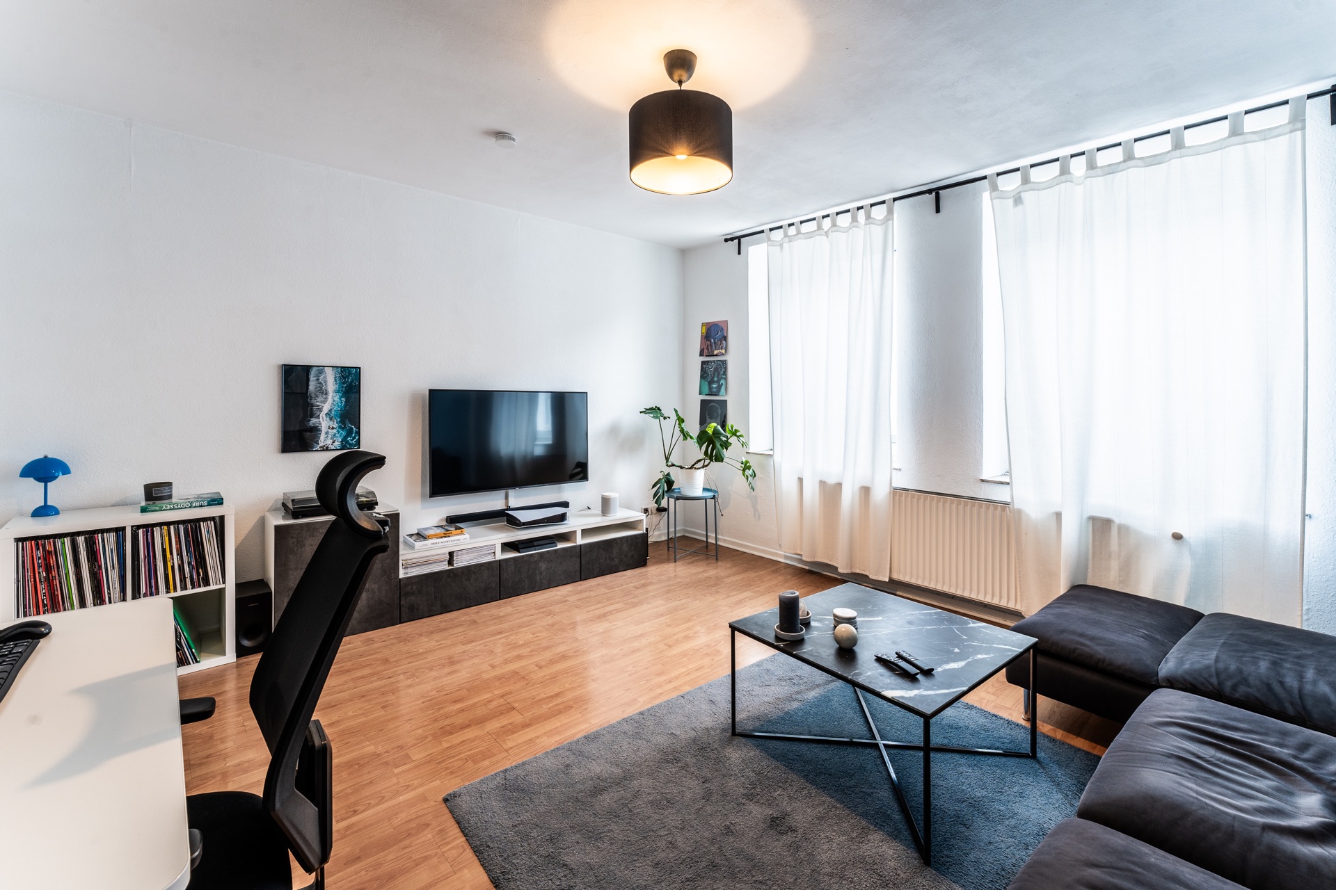 Modernes Wohnzimmer mit Sofa, Couchtisch, Fernseher, Schreibtisch und großen Fenstern mit weißen Vorhängen.