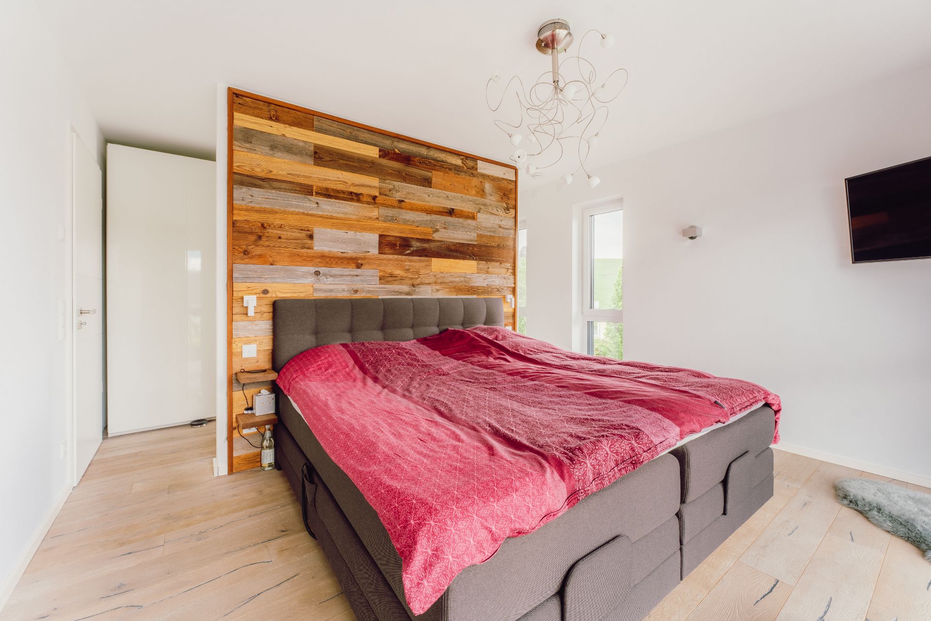 Modernes Schlafzimmer mit grauem Bett und roter Decke vor Holzwand.