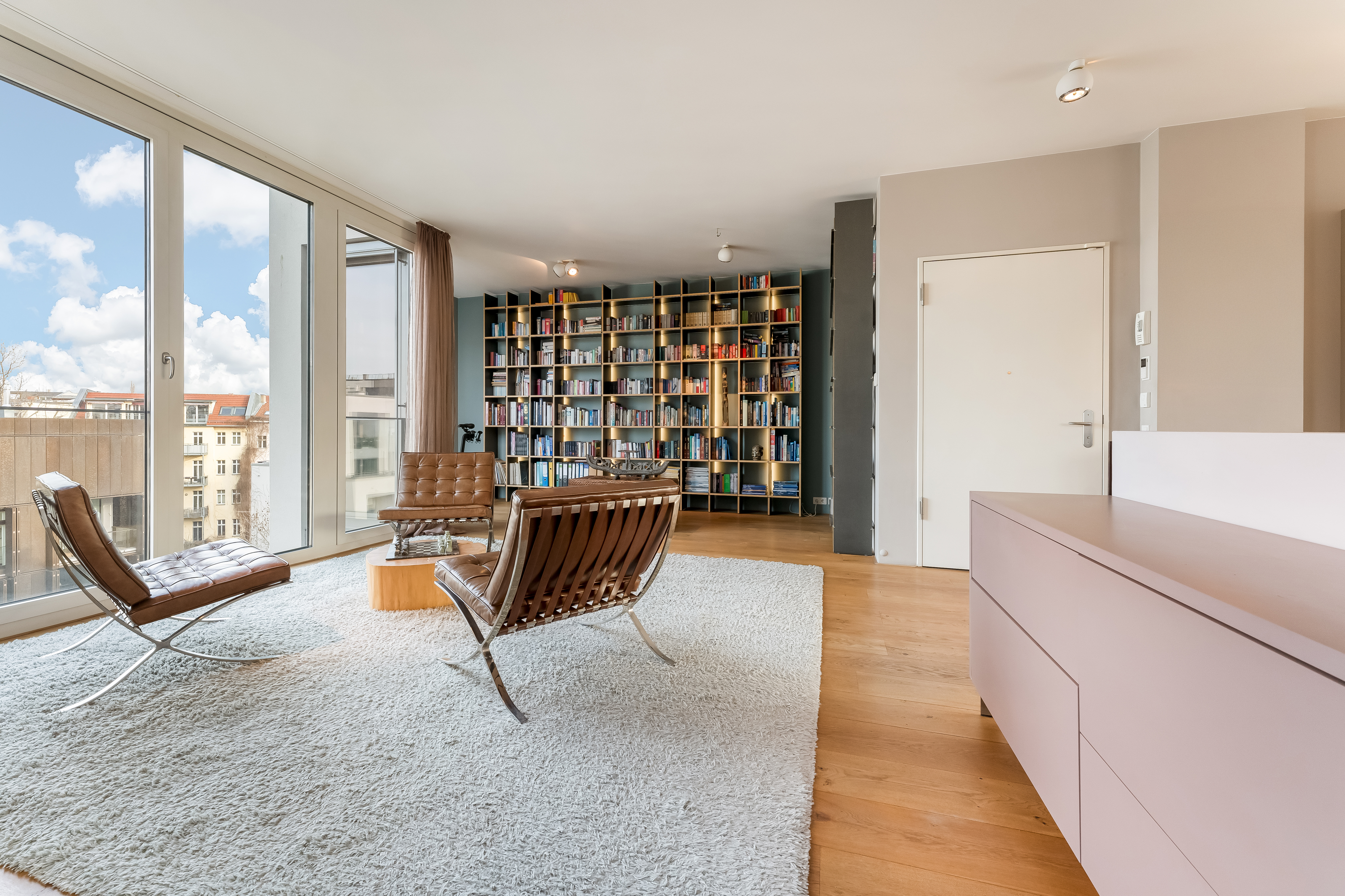 Helles Wohnzimmer mit großen Fenstern, zwei Ledersesseln, Teppich und Bücherregal.