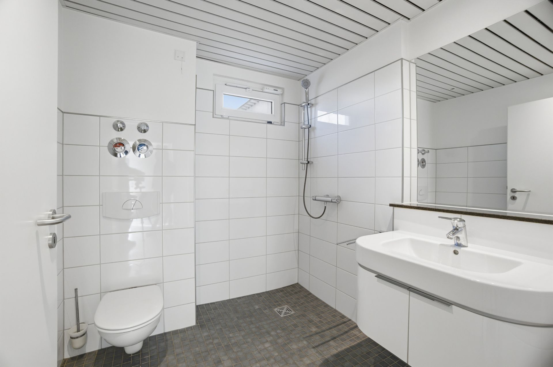 Modernes Bad mit ebenerdiger Dusche, WC und Waschbecken, weiße Fliesen an Wand und Decke.