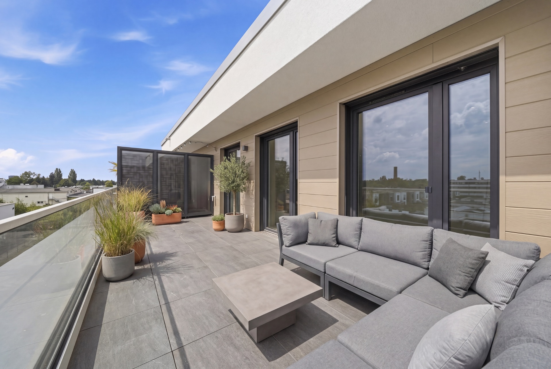 Große Terrasse mit grauem Sofa, Tisch und mehreren Pflanzen in Töpfen neben Glasgeländer.