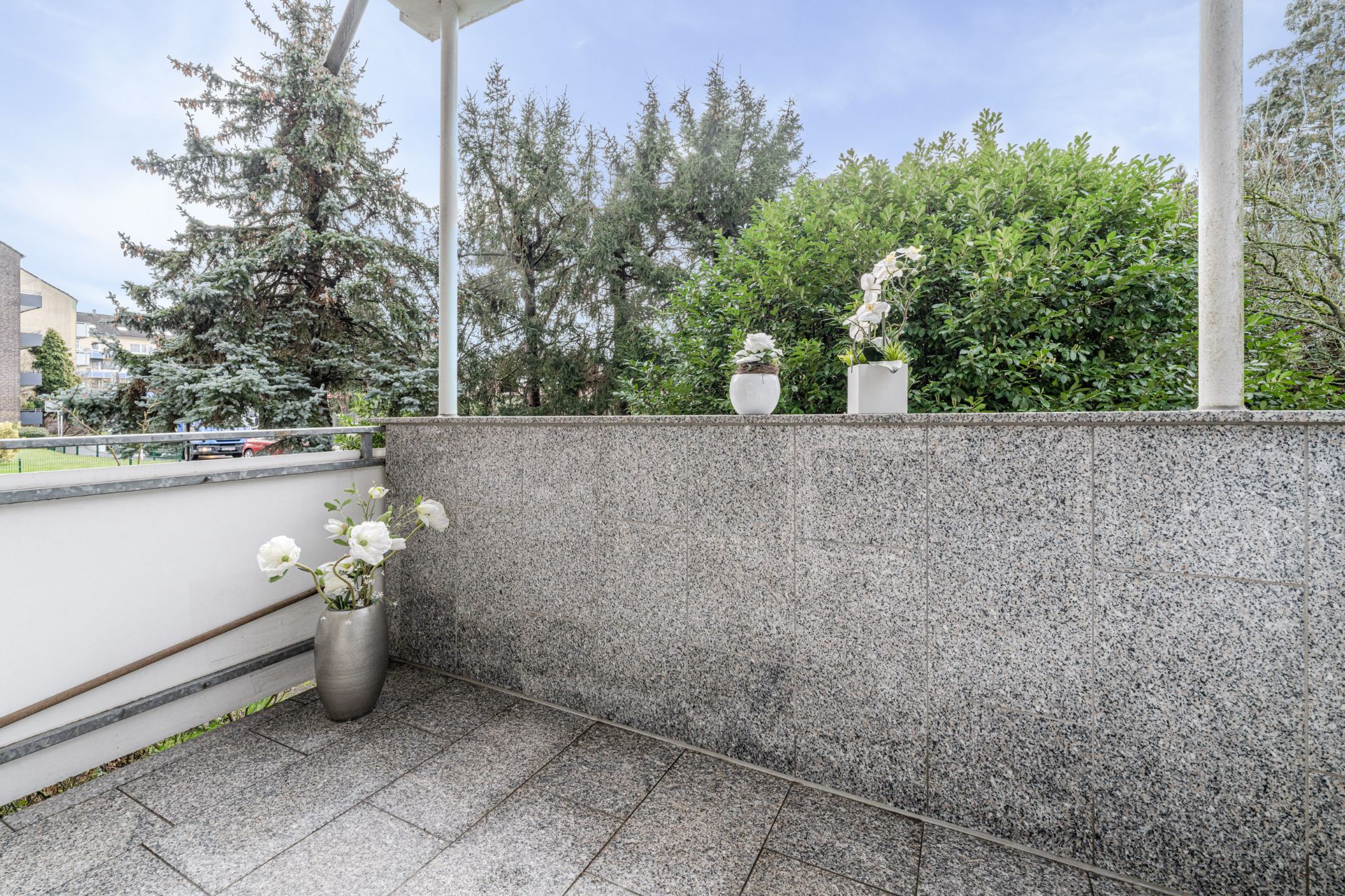Balkon mit grauen Fliesen, zwei Blumentöpfen auf der Mauer und einem Blumentopf in der Ecke.