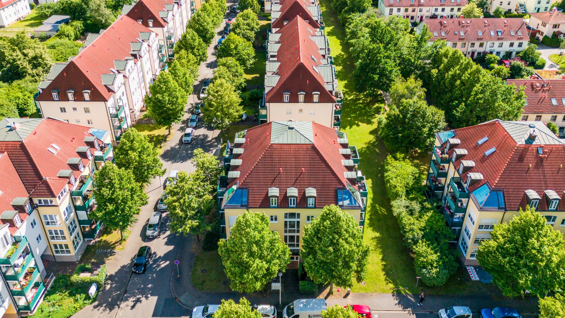 Luftaufnahme von Wohngebäuden mit roten Dächern, Bäumen und parkenden Autos an der Straße.