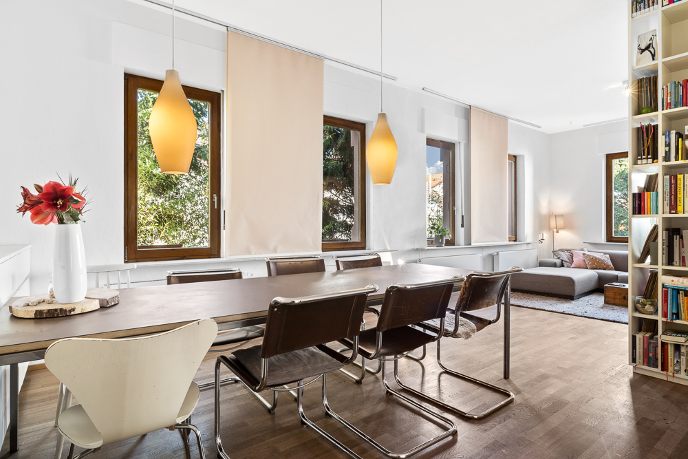 Essbereich mit großem Tisch, Stühlen, Hängelampen und Wohnzimmer mit Sofa im Hintergrund.