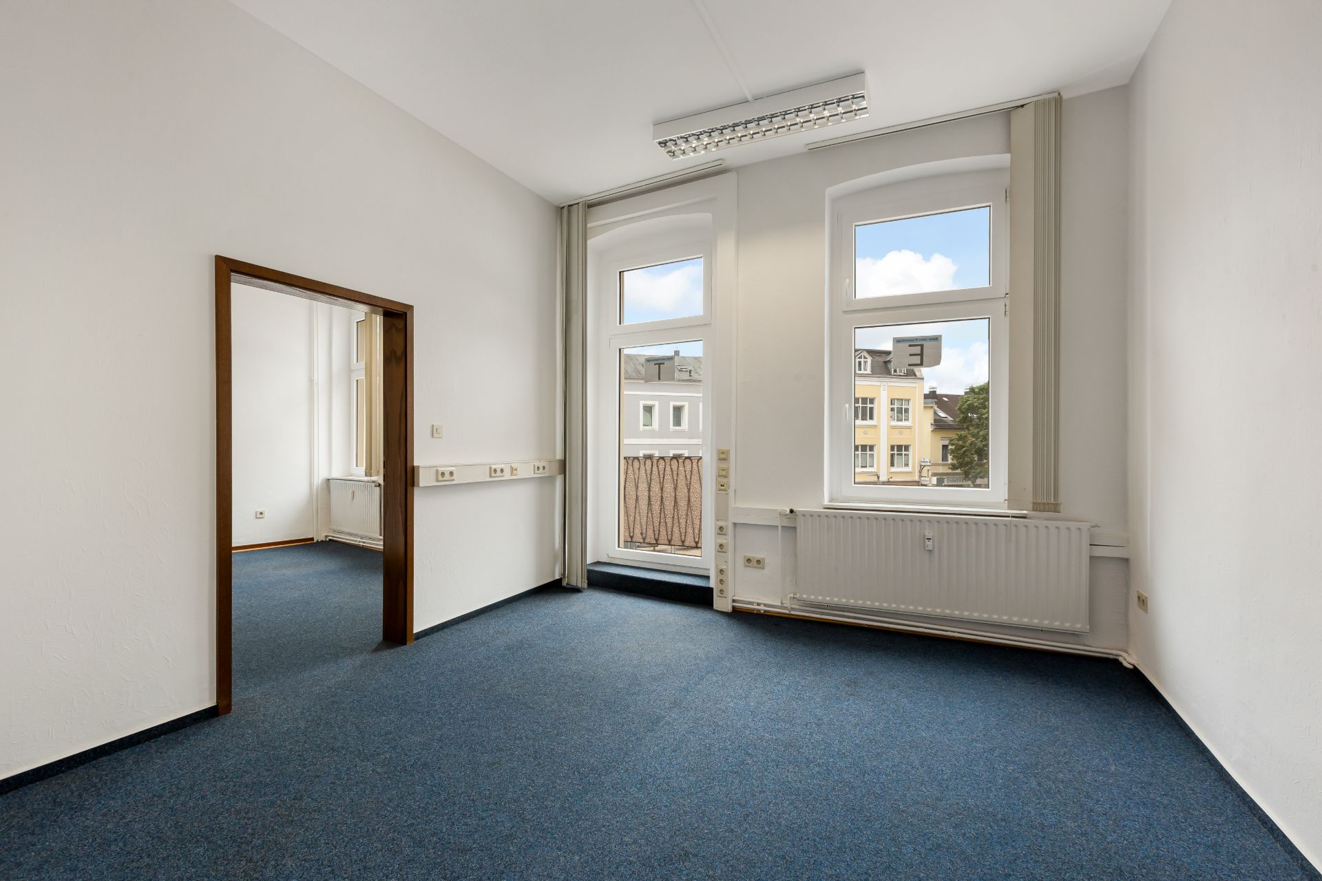 Helles Zimmer mit blauem Teppich, zwei Fenstern, Heizung und Durchgang zu einem weiteren Raum.