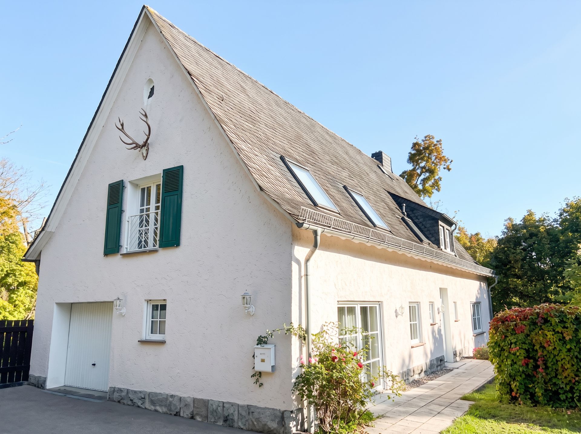 Weißes Einfamilienhaus mit steilem Dach, Garage und grünen Fensterläden, umgeben von Garten.