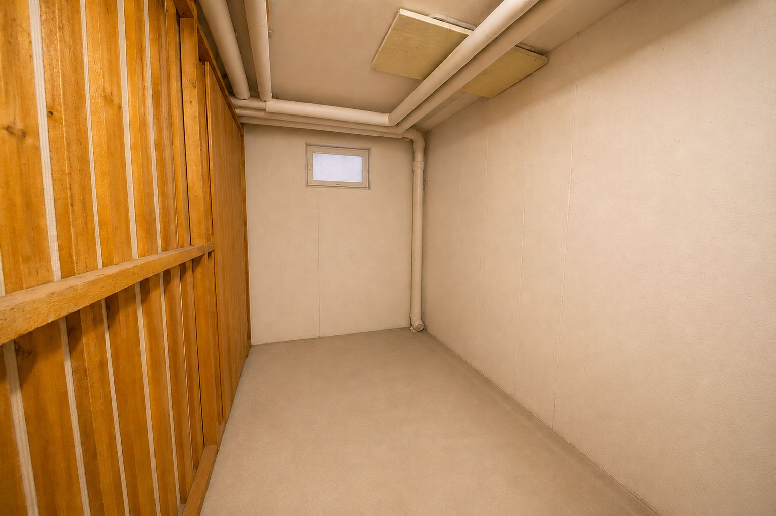 Kleiner, leerer Raum mit kleinem Fenster, sichtbaren Rohren und Holzwand auf der linken Seite.