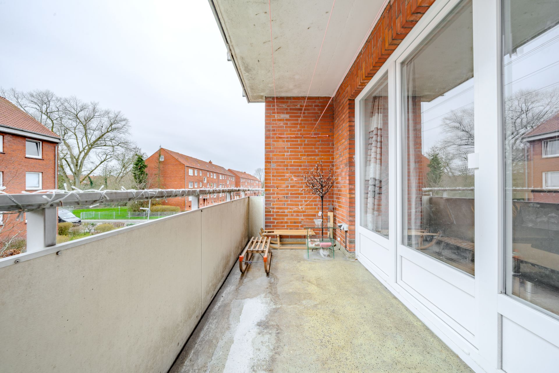 Balkon mit Bank, Blumentopf und Glasschiebetür, Blick auf rote Backsteingebäude.