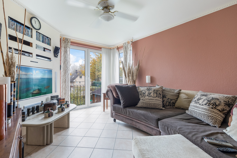 Helles Wohnzimmer mit Ecksofa, Kissen, Couchtisch und großen Fenstern mit Blick nach draußen.