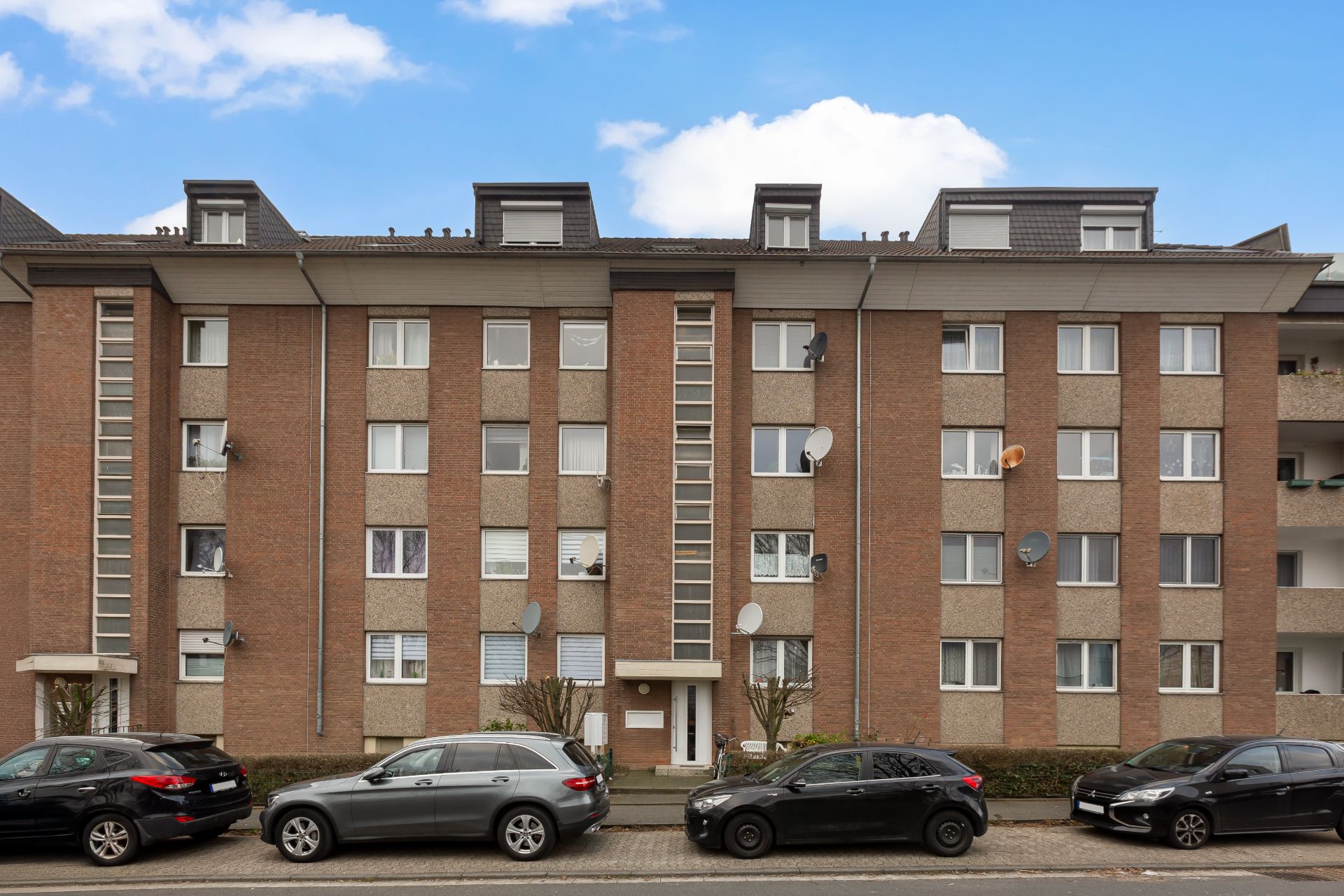 Mehrfamilienhaus mit Backsteinfassade, mehreren Fenstern und parkenden Autos davor.