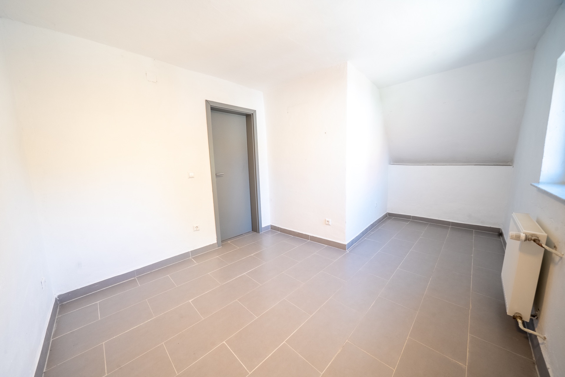 Heller, leerer Raum mit grauen Fliesen, weißer Wand, Tür und Fenster.