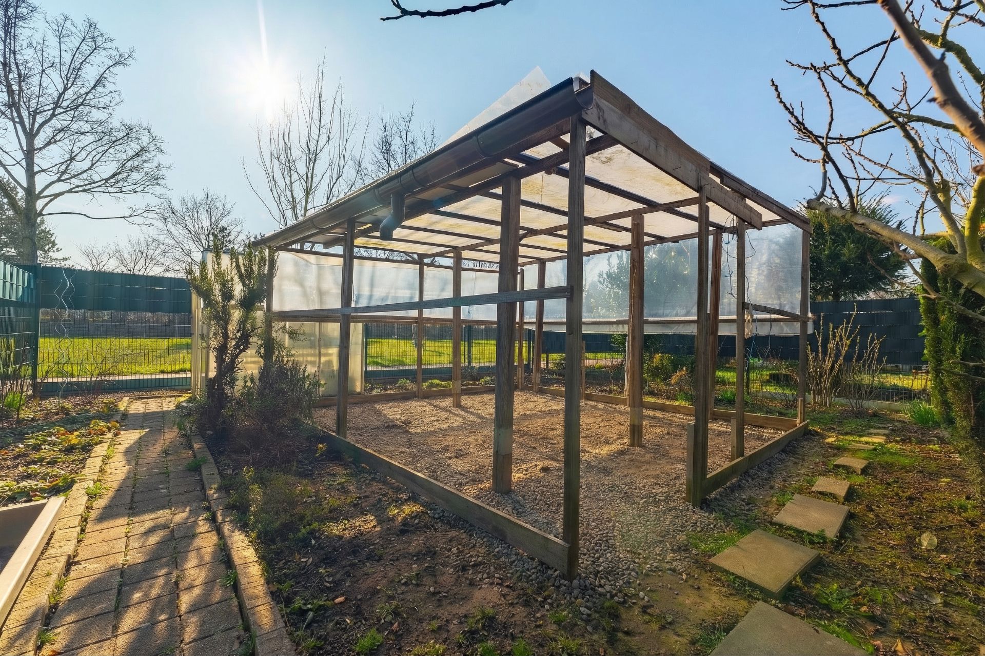 Ein leerer Gewächshausrahmen aus Holz mit transparenten Wänden im Garten bei Sonnenschein.
