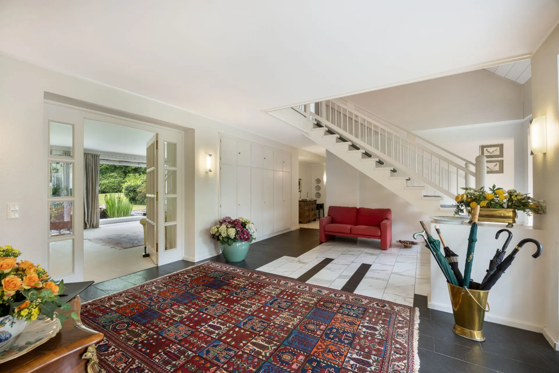 Heller Eingangsbereich mit Teppich, rotem Sofa, Treppe und großem Blumenstrauß.