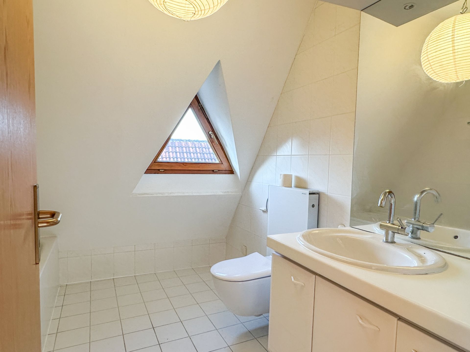 Helles Badezimmer mit Dreiecksfenster, Waschbecken, Spiegel und WC.