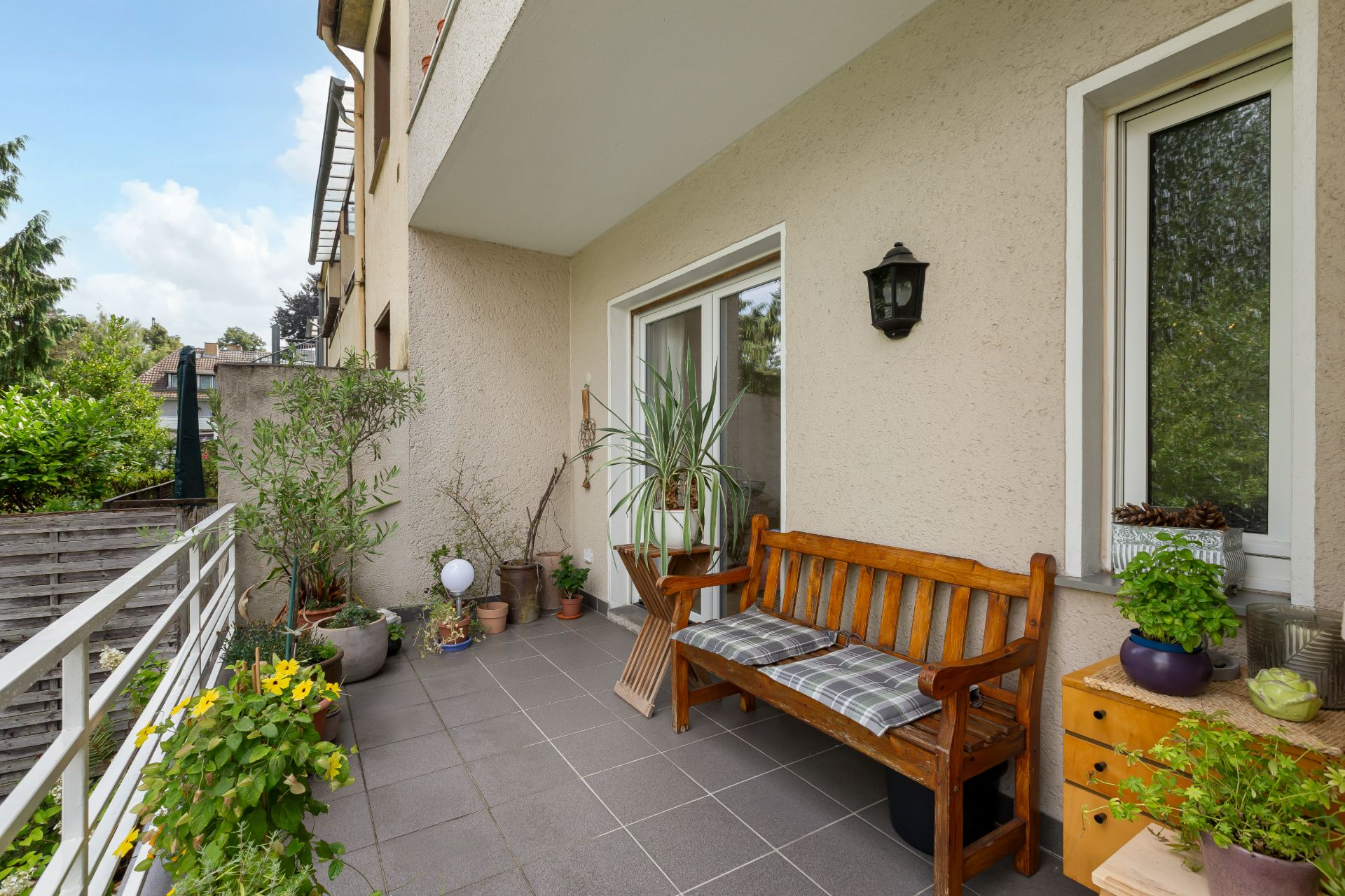 Balkon mit Holzbänken, Pflanzen, kleiner Kommode und Zugangstür, umgeben von Grün.