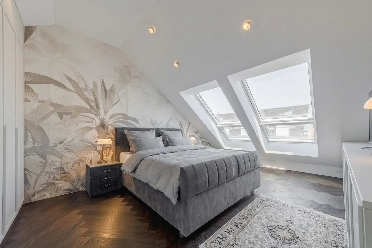 Helles Schlafzimmer mit Doppelbett, zwei Dachfenstern und einem Wandbild mit Pflanzenmotiv.