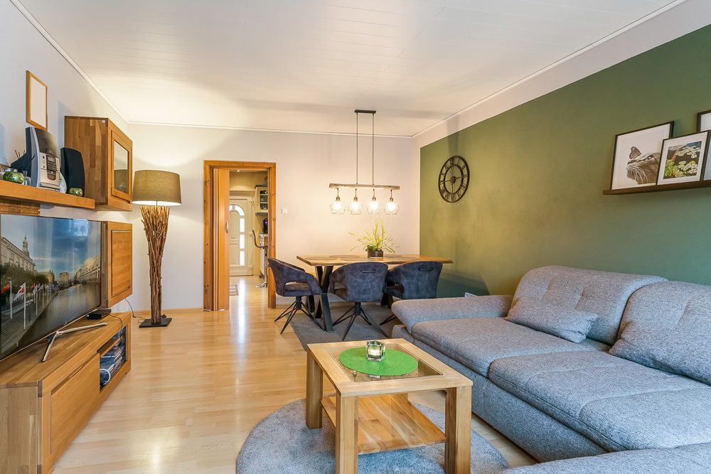 Wohnzimmer mit grauem Sofa, Holzmöbeln, Esstisch, Fernseher und grüner Wand.