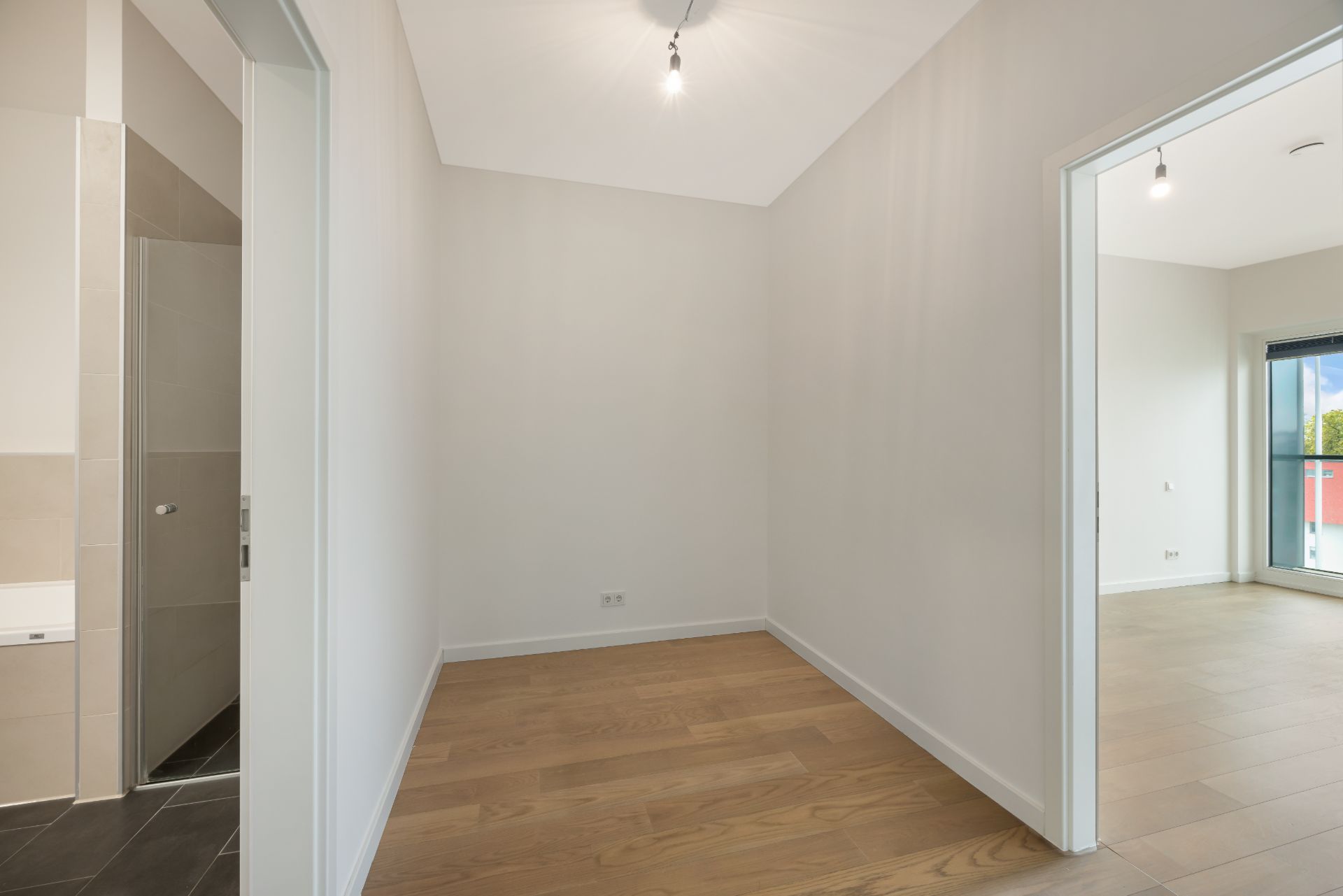 Leerer Raum mit Holzfußboden, zwei offenen Türen, sichtbarem Bad und angrenzendem Wohnzimmer.
