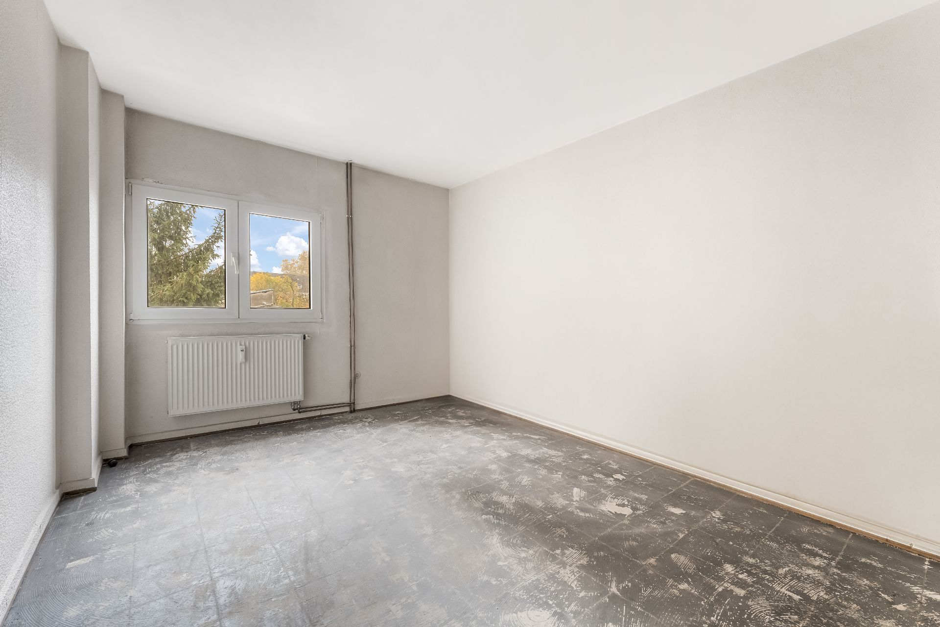 Leeres Zimmer mit grauem Boden, weißen Wänden und Fenster mit Blick ins Grüne.