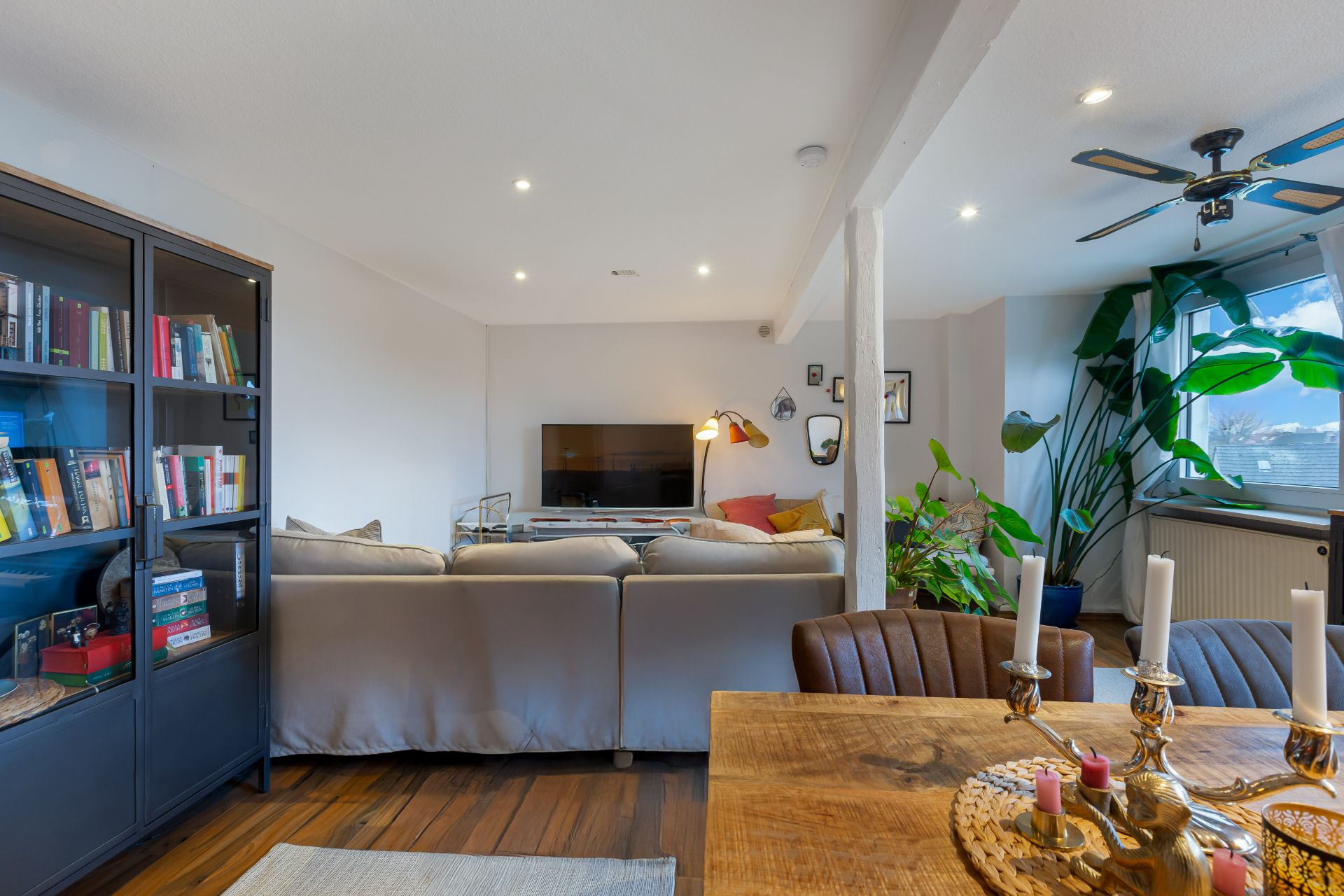 Helles Wohnzimmer mit Sofa, Fernseher, Bücherregal, Holztisch, Pflanzen und großen Fenstern.
