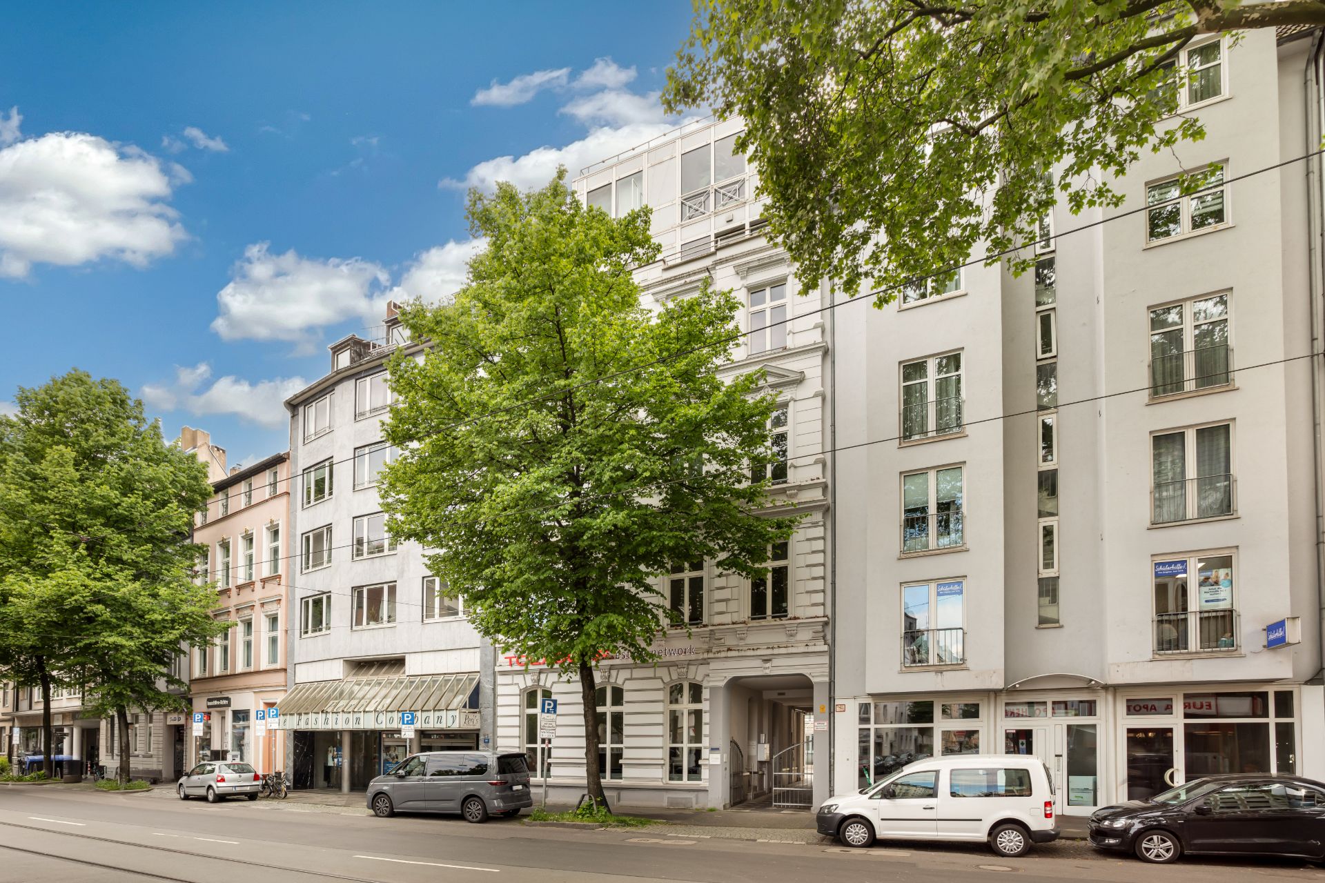 Mehrstöckige Wohngebäude an einer Straße mit geparkten Autos und Bäumen im Vordergrund.