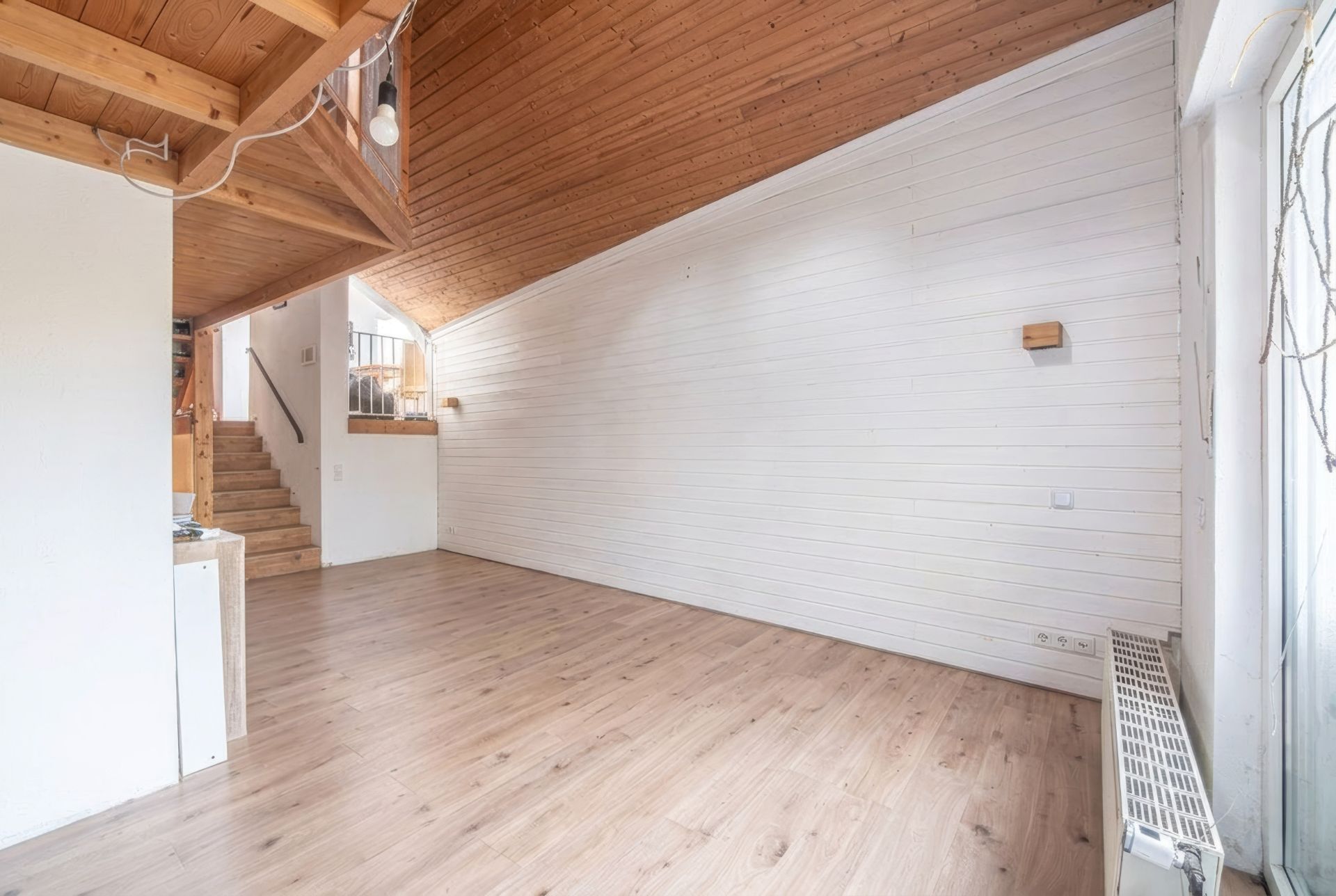 Heller Raum mit Holzdecke, weißer Holzvertäfelung, Holzboden und Treppe im Hintergrund.