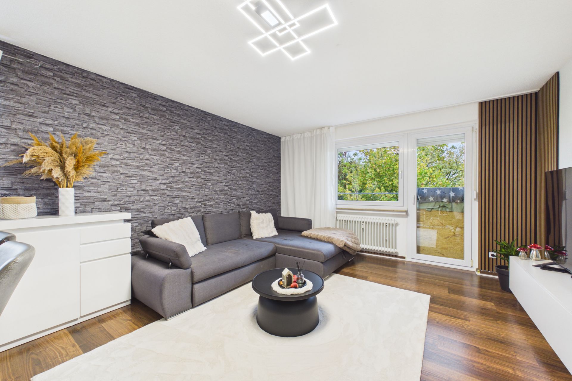 Modernes Wohnzimmer mit grauer Eckcouch, Teppich, Couchtisch und großer Fensterfront zum Balkon.