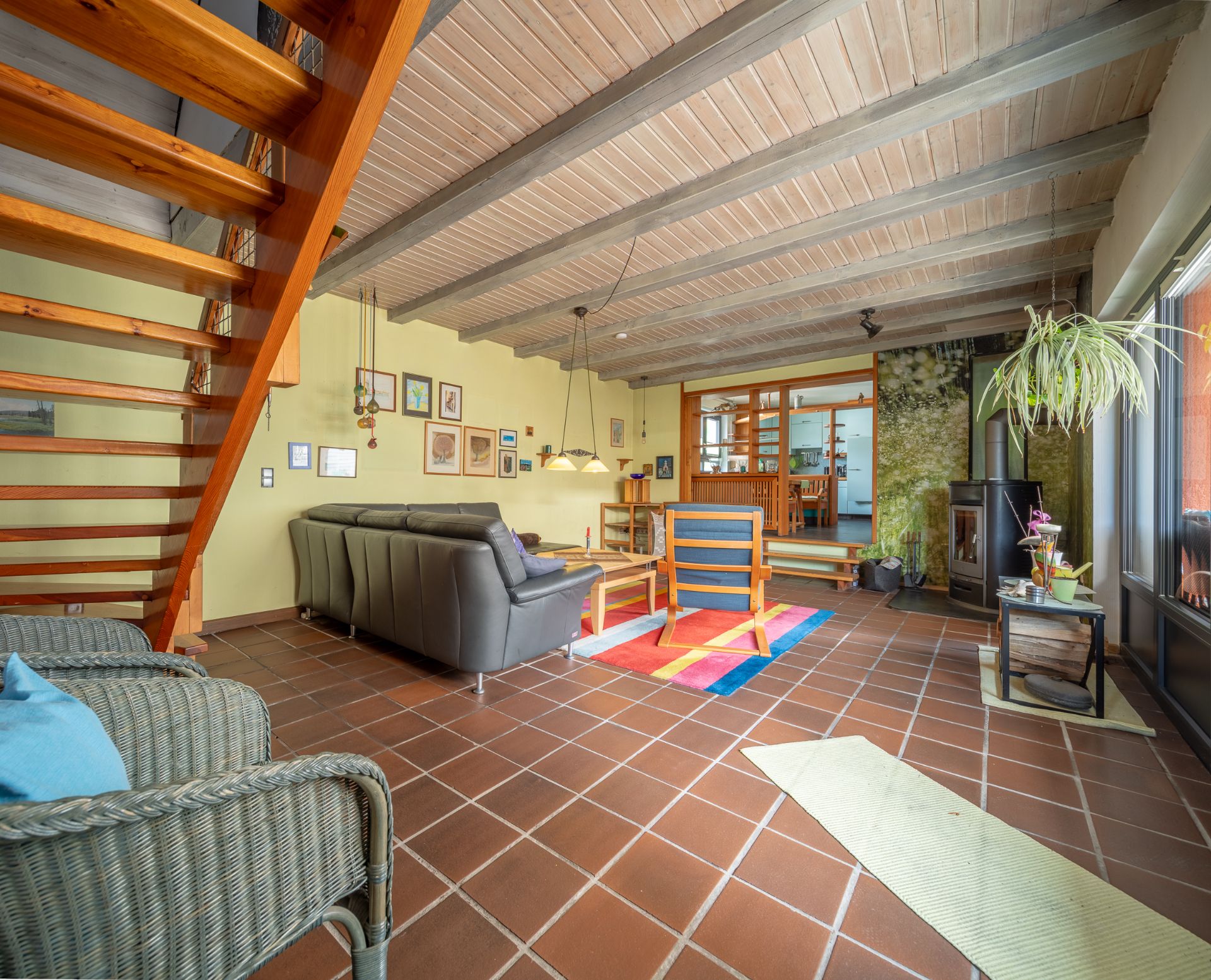 Wohnzimmer mit braunen Fliesen, Holzdecke, Sofa, Sessel, Kamin und Holztreppe.