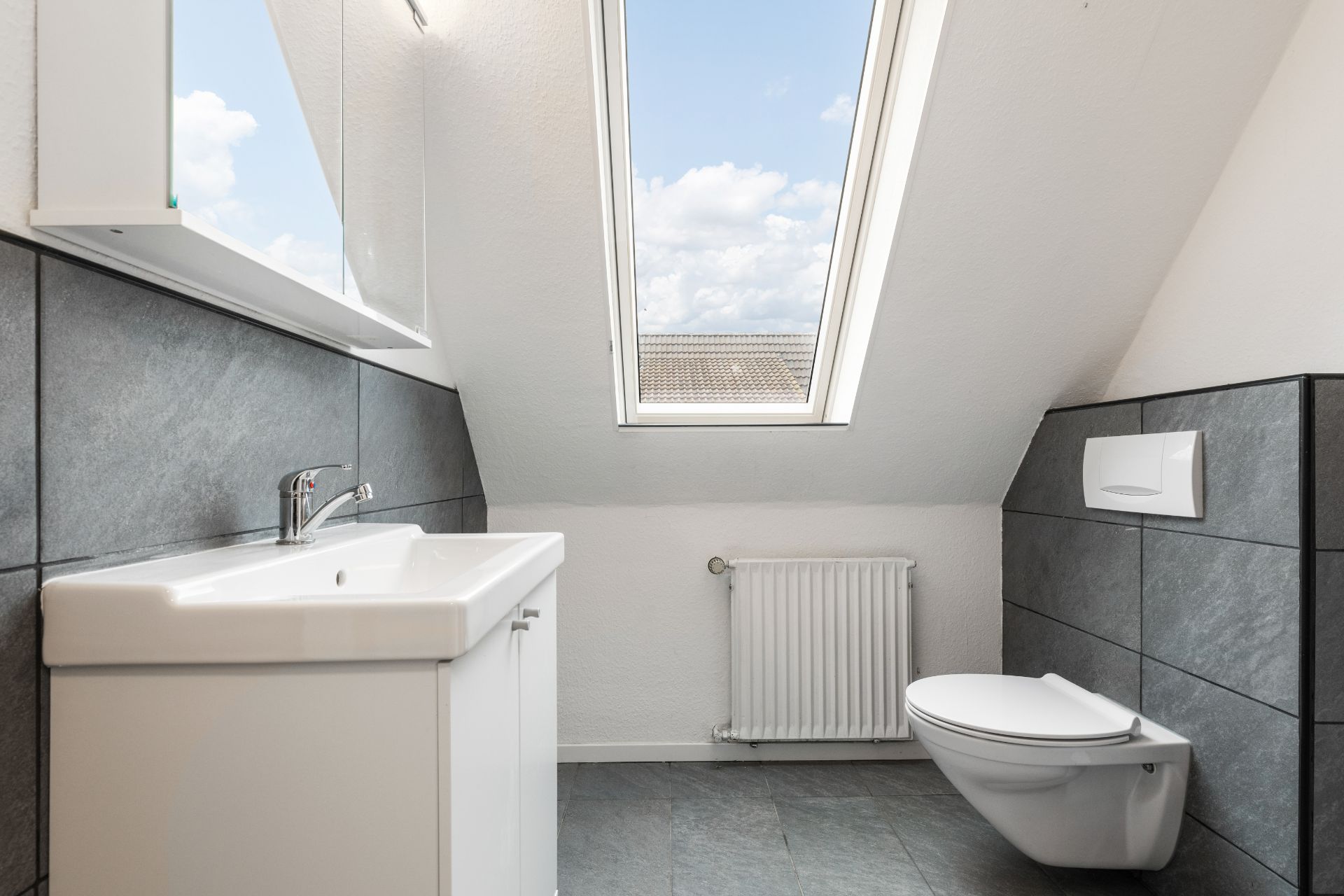 Modernes Bad mit Waschbecken, Wand-WC und Dachfenster, graue Fliesen an Wänden und Boden.