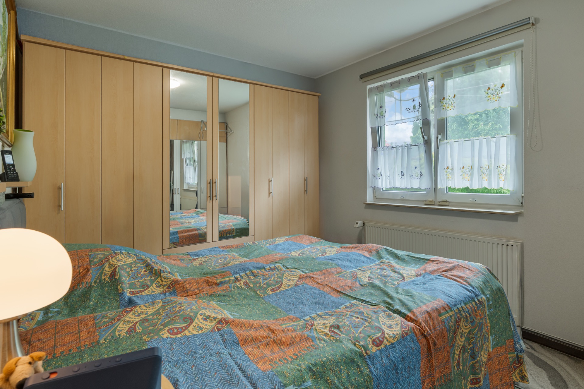 Schlafzimmer mit großem Kleiderschrank, Doppelbett und Fenster mit Gardinen.
