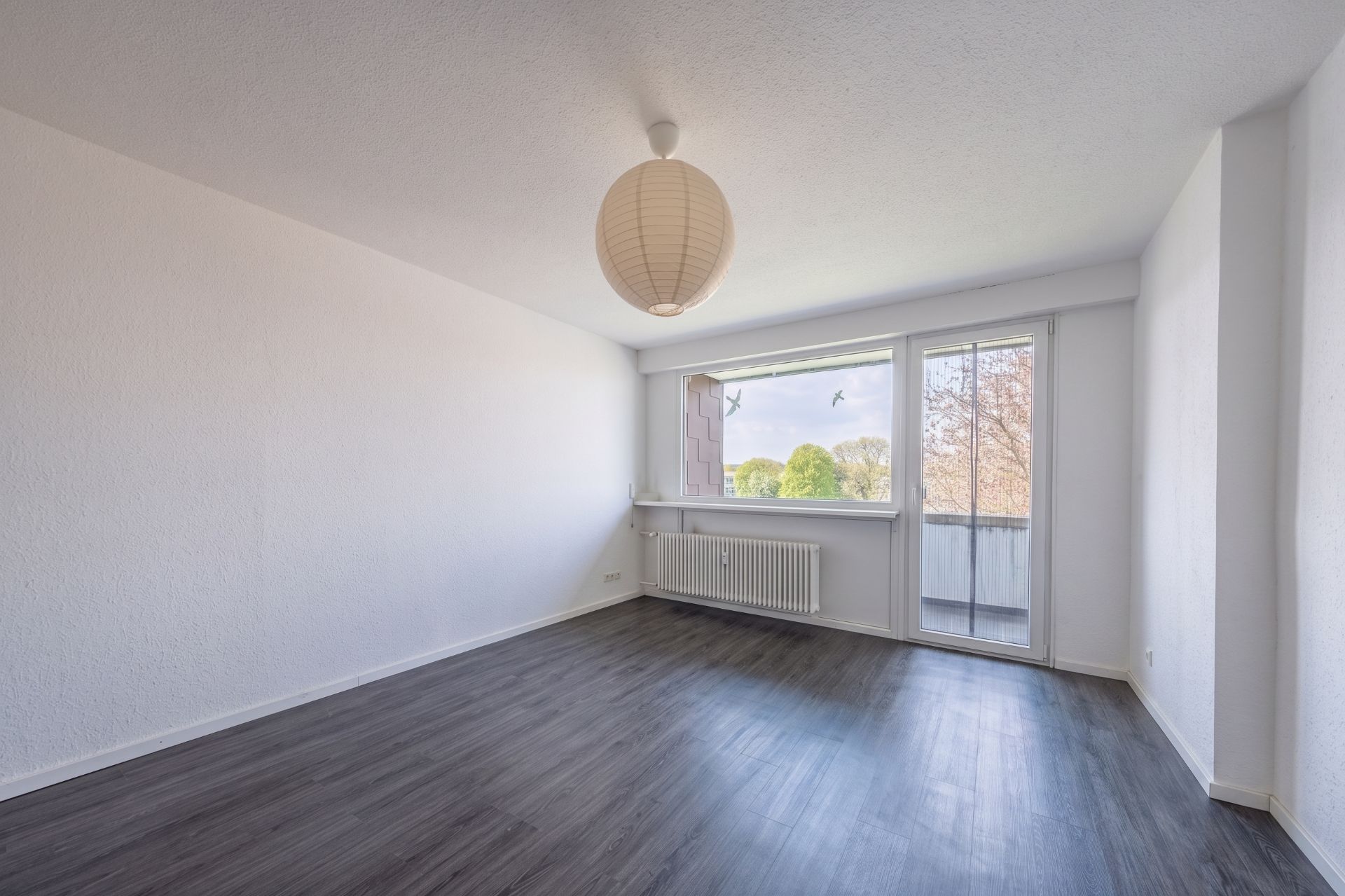 Leeres Zimmer mit dunklem Boden, großem Fenster, Glastür und Lampe an der Decke.