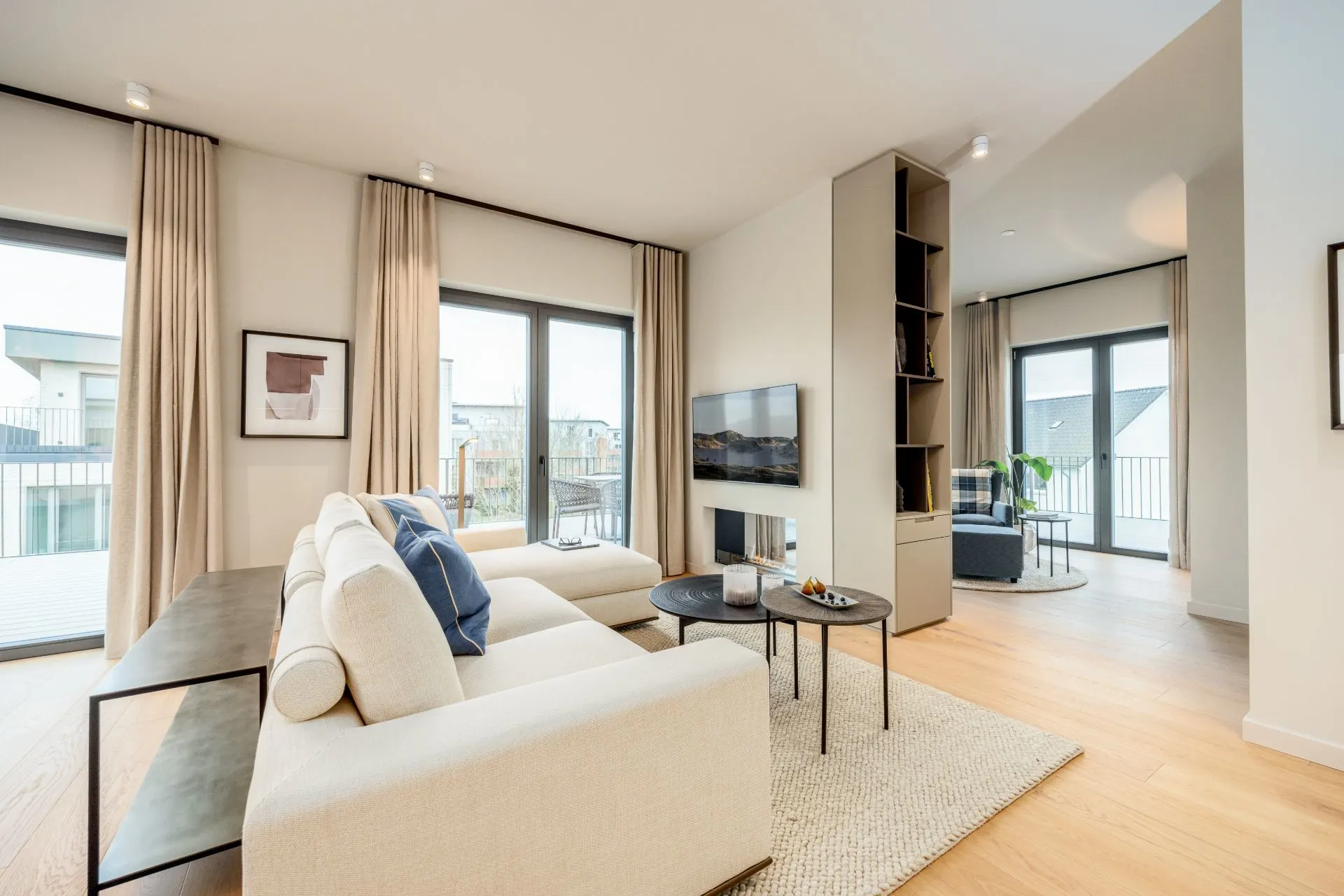 Helles Wohnzimmer mit Sofa, Couchtischen, TV und großen Fenstern mit Vorhängen.