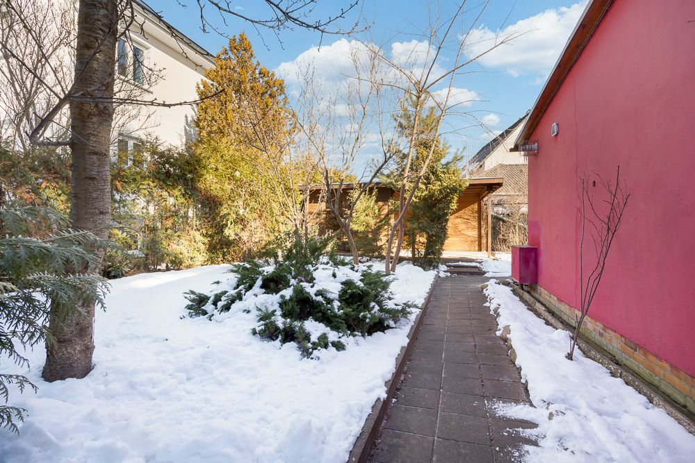 Gepflasterter Weg mit Schnee im Garten, rechts ein rotes Gebäude, Sträucher und Bäume im Hintergrund.