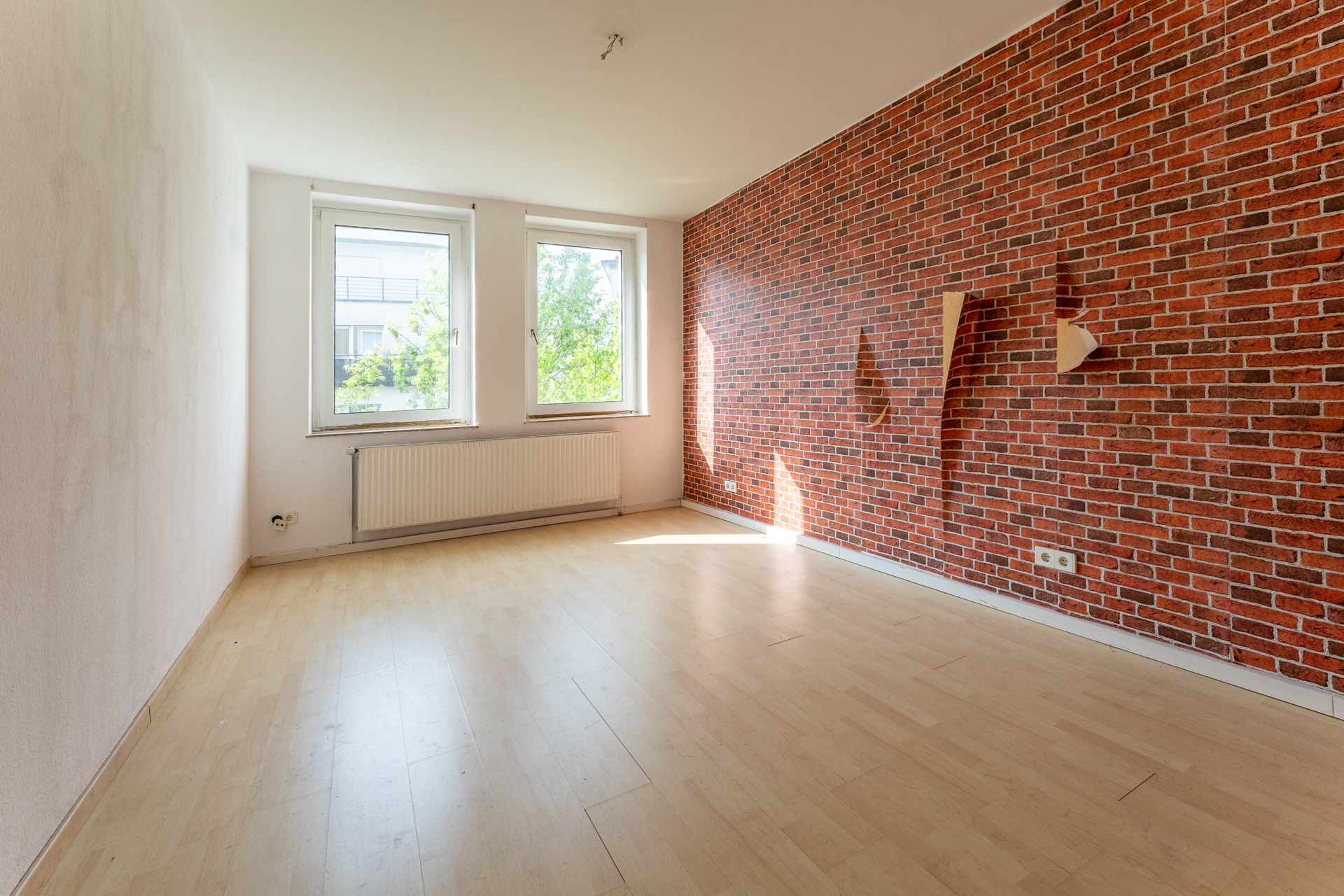 Leerer Raum mit heller Holzboden, zwei Fenstern und einer roten Backsteinwand.