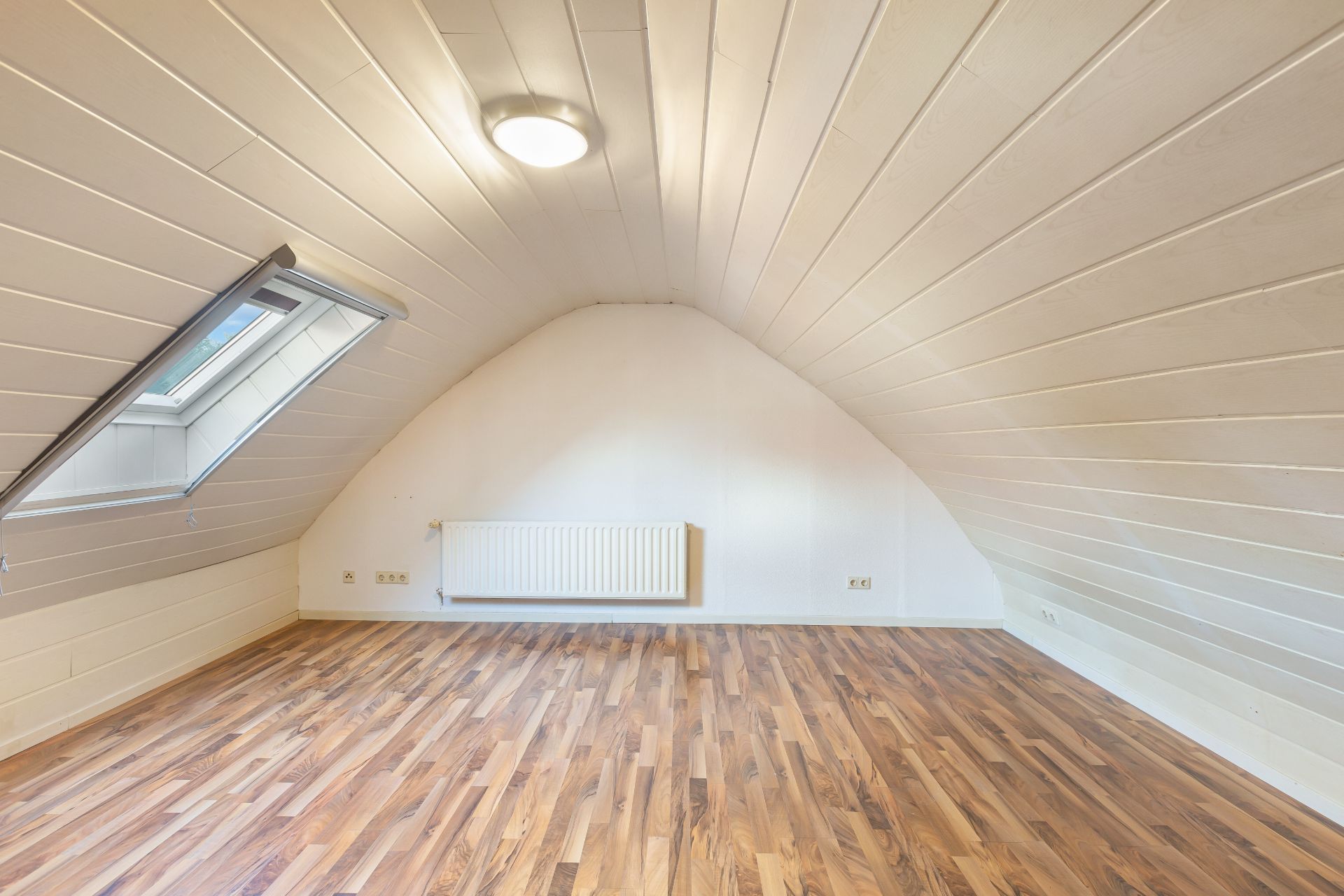 Helles Dachzimmer mit Holzfußboden, Dachfenster und Heizkörper an der Wand.