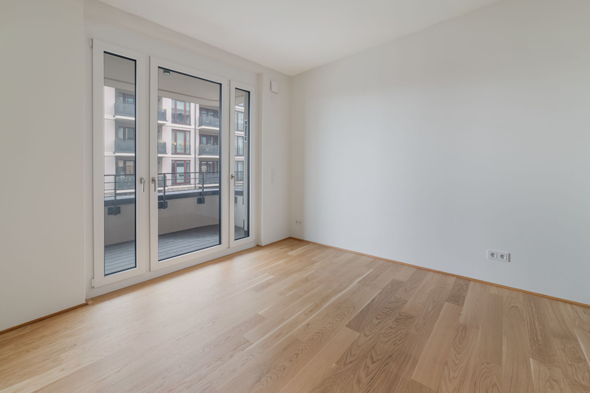 Helles Zimmer mit Parkettboden und großer Glastür zu einem Balkon, moderne Wohnanlage im Hintergrund.