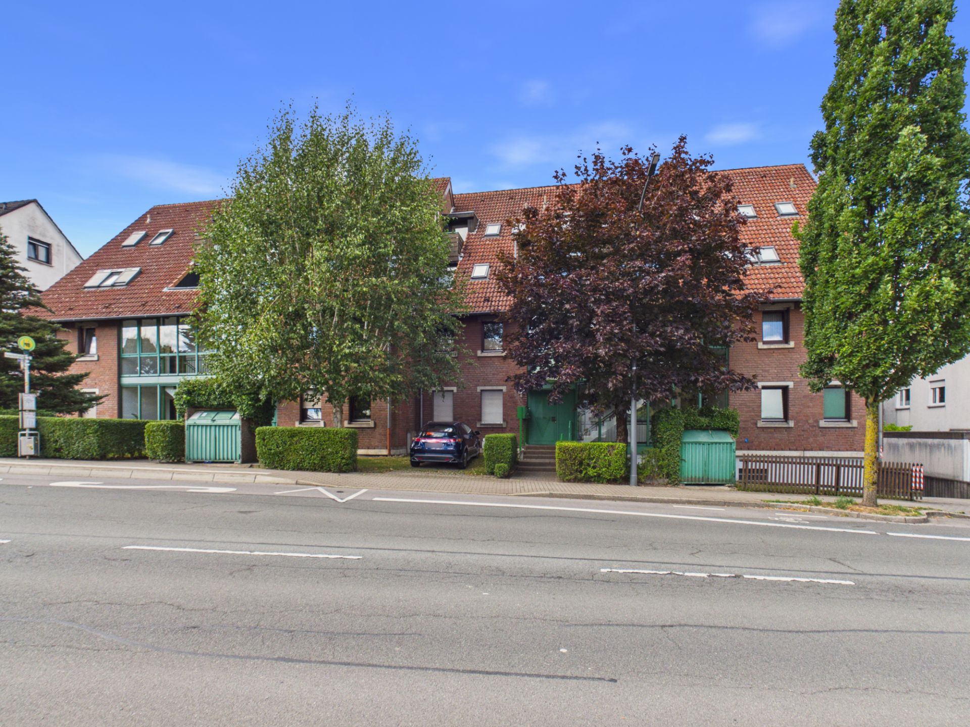 Mehrfamilienhaus mit rotem Dach, Bäumen davor und geparktem Auto an einer Straße.