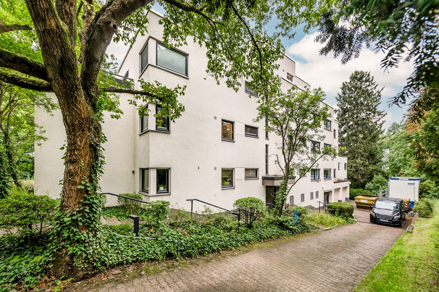 Mehrfamilienhaus mit weißer Fassade, großen Fenstern und umgeben von Bäumen und Grünflächen.