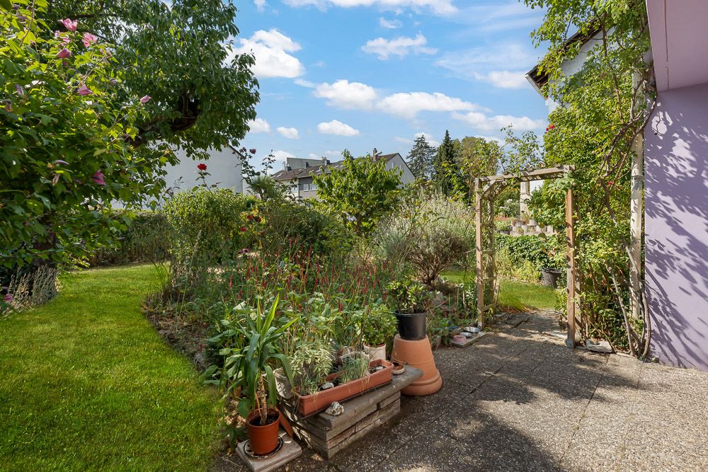 Garten mit Rasenfläche, Blumenbeeten, Pflanzkübeln und kleiner Terrasse neben einem Haus.