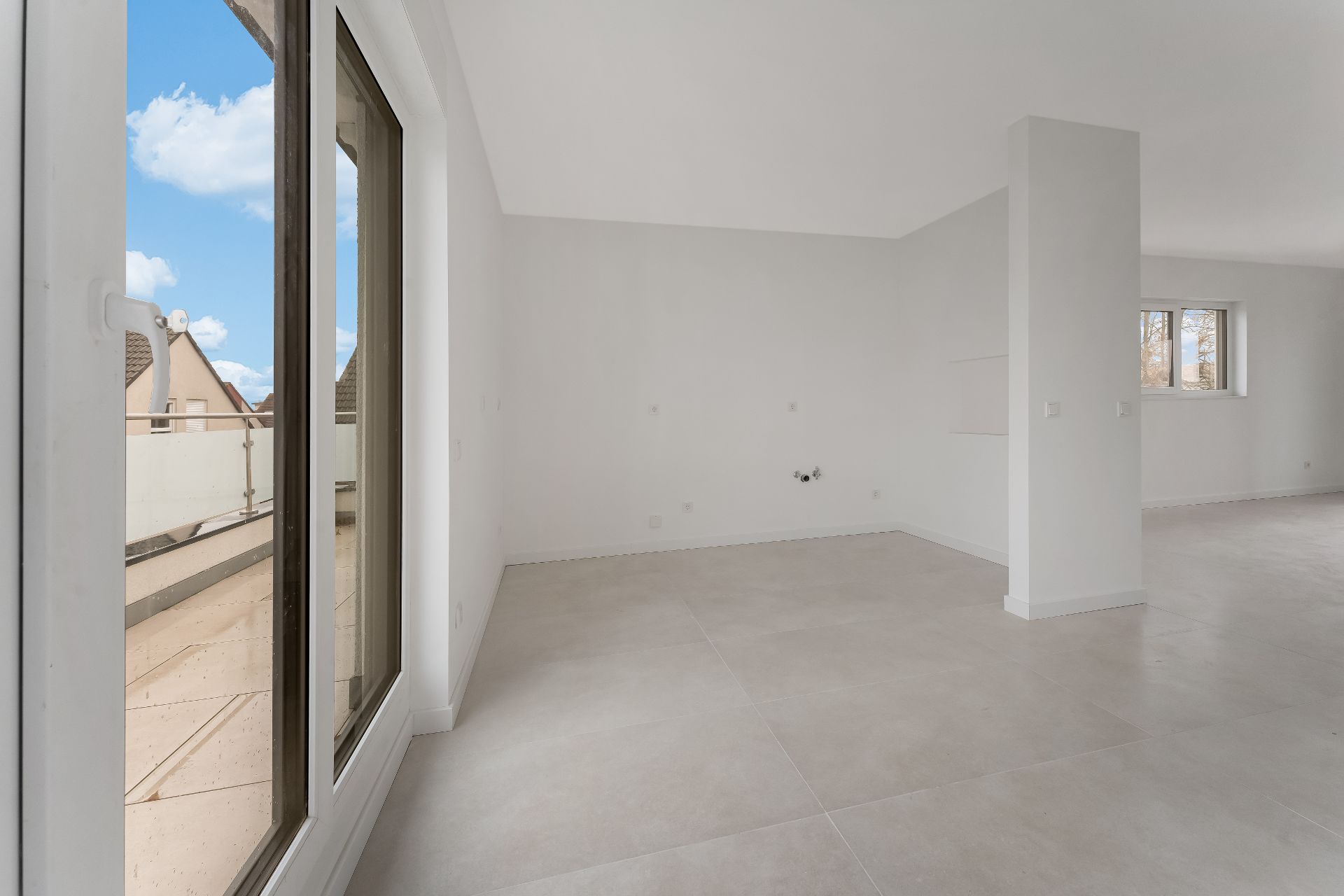 Helles, leeres Zimmer mit großen Fliesen und Zugang zu einem Balkon durch eine Glastür.
