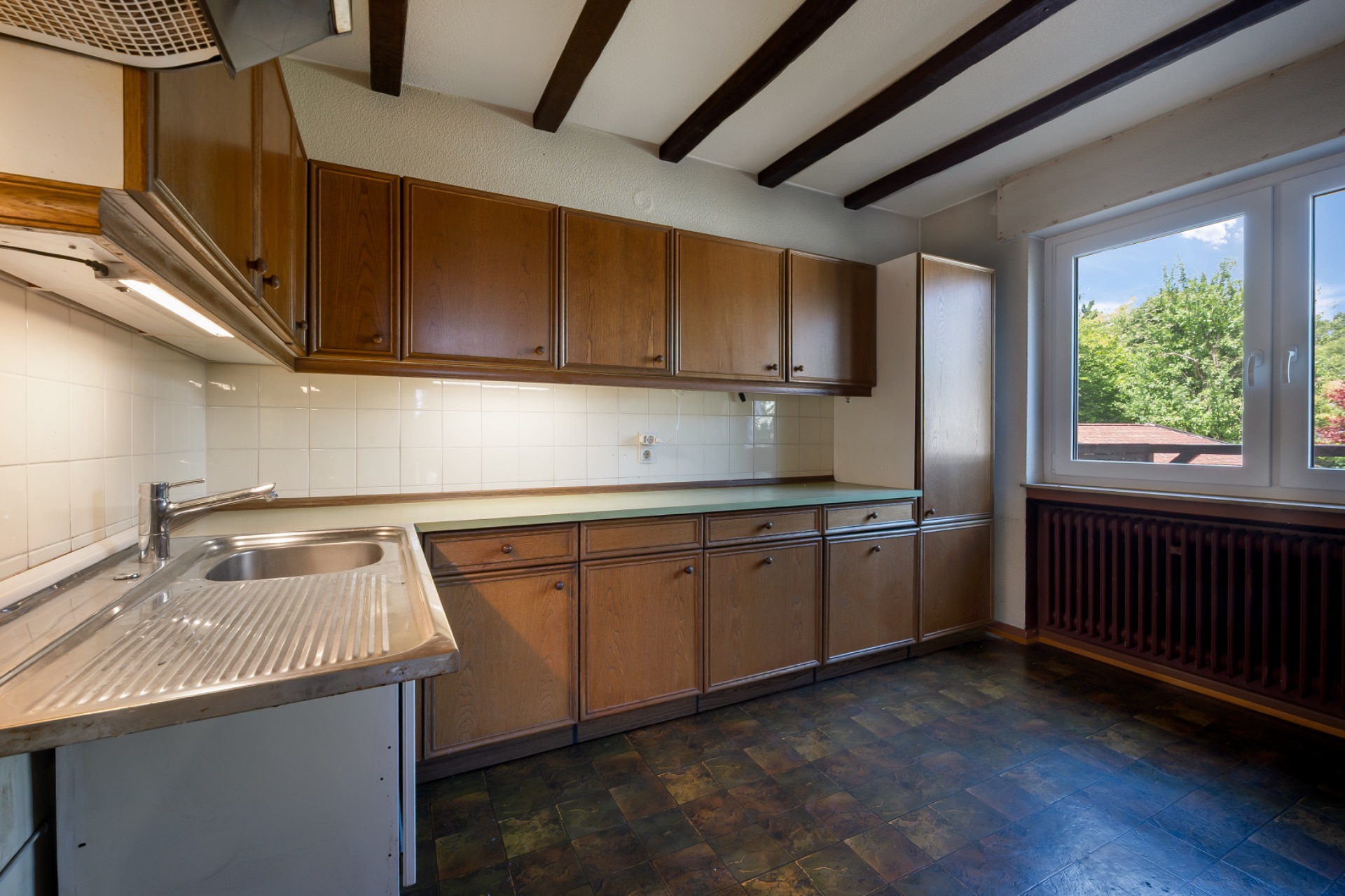 Küche mit braunen Holzschränken, Spüle, Fliesenwand und großem Fenster mit Blick ins Grüne.