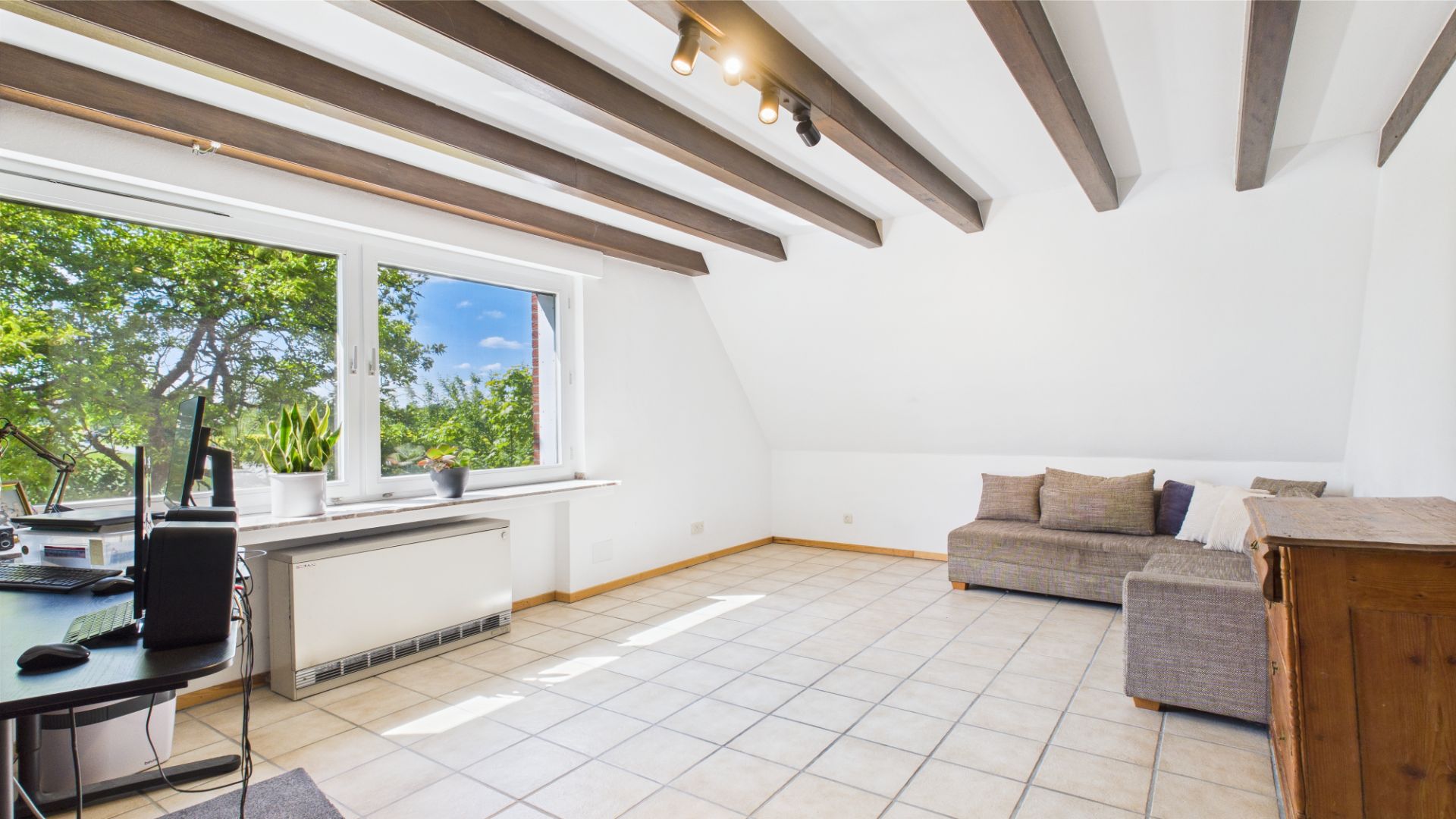 Helles Zimmer mit Fliesenboden, Schreibtisch, Fenster, grauem Sofa und Holzbalken an der Decke.