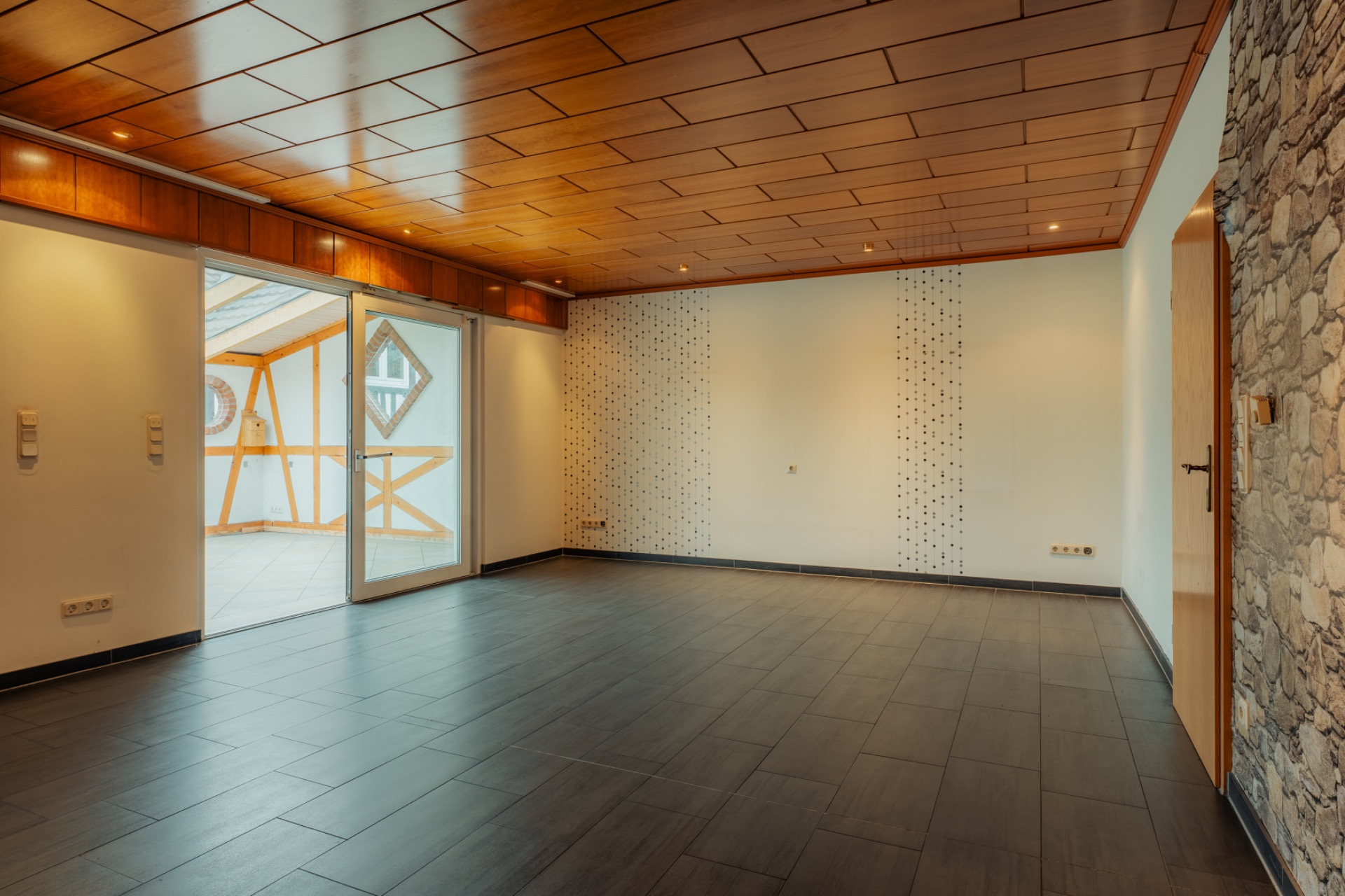Leeres Wohnzimmer mit Holzboden, Holzdecke, Steintapete und Zugang zu verglastem Balkon.