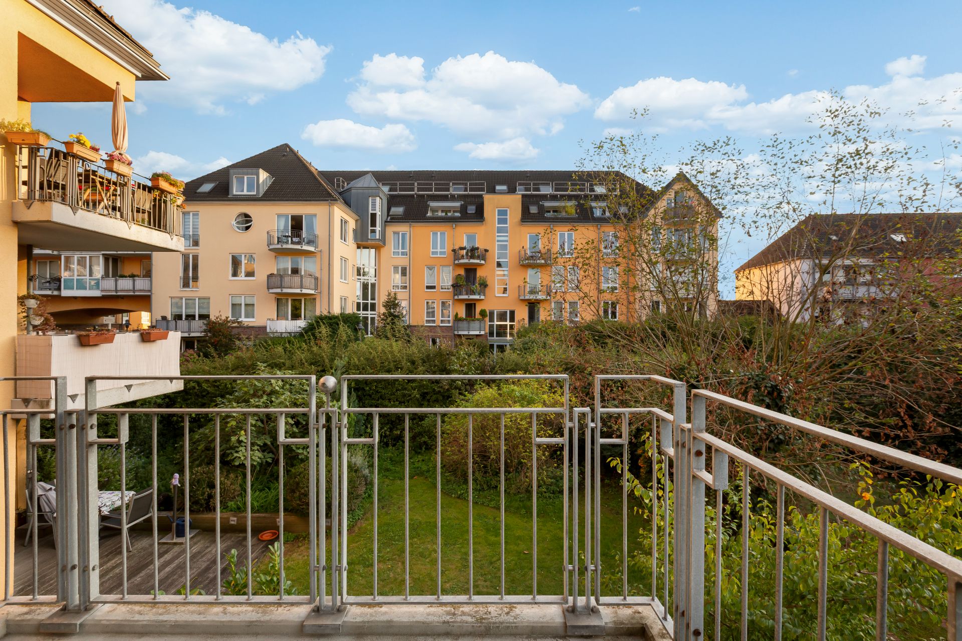 Blick von einem Balkon auf einen grünen Garten und gelbe Wohngebäude im Hintergrund.