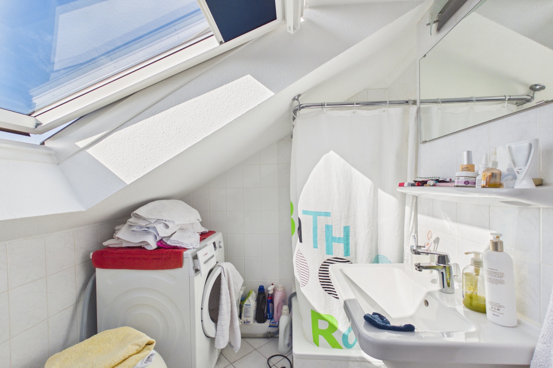 Helles Badezimmer mit Dachfenster, Waschmaschine, Waschbecken und Duschvorhang.