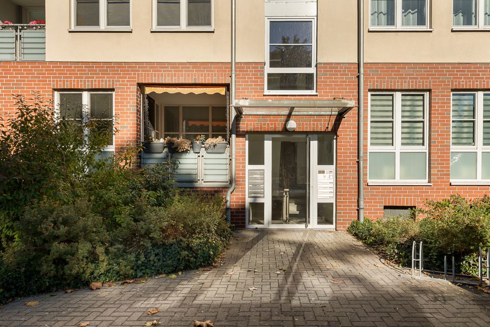 Eingang eines Mehrfamilienhauses mit Glasdach, Briefkästen und bepflanztem Vorgarten.