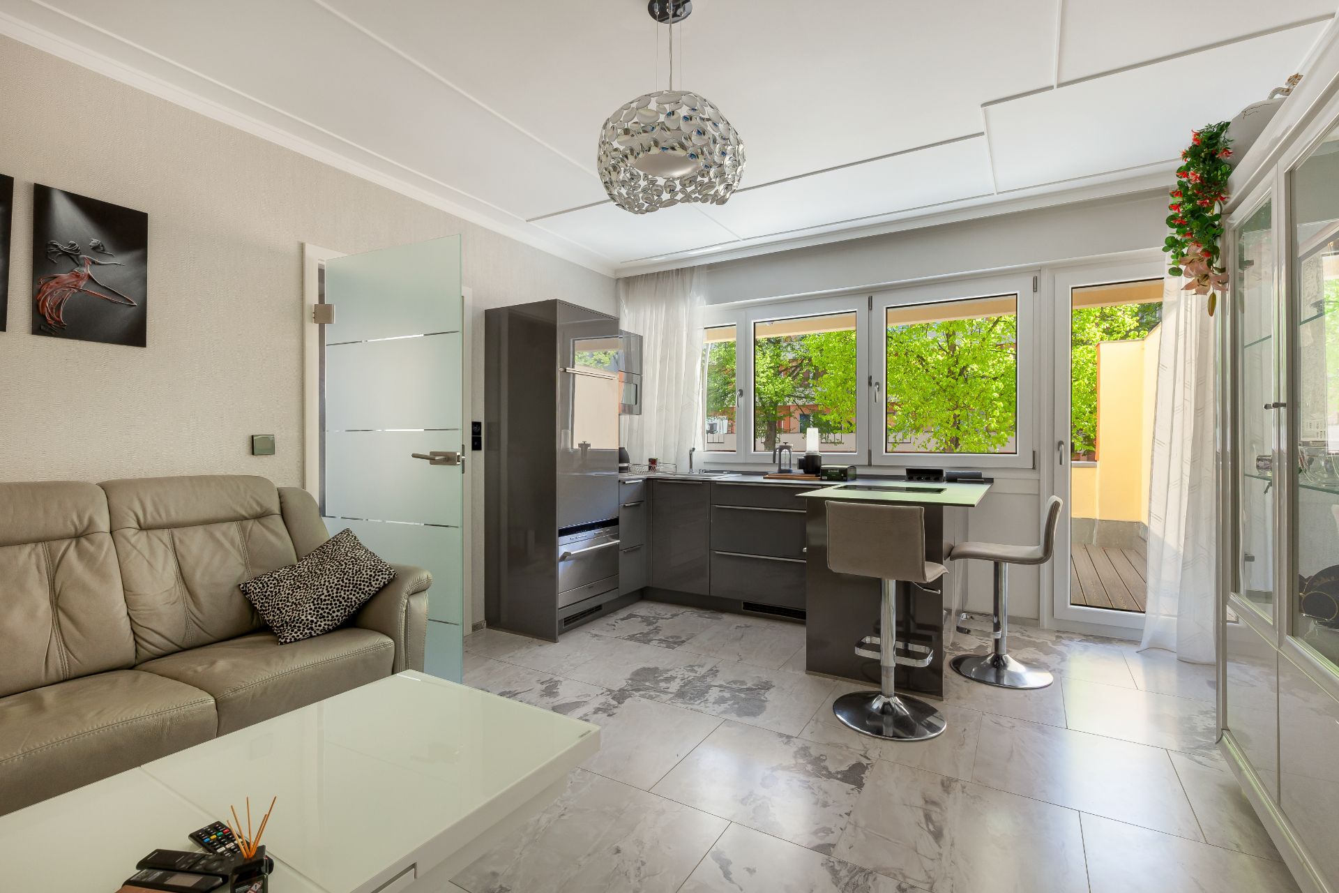 Modernes Wohnzimmer mit Sofa, Glastisch, offener Küche und großen Fenstern mit Blick ins Grüne.